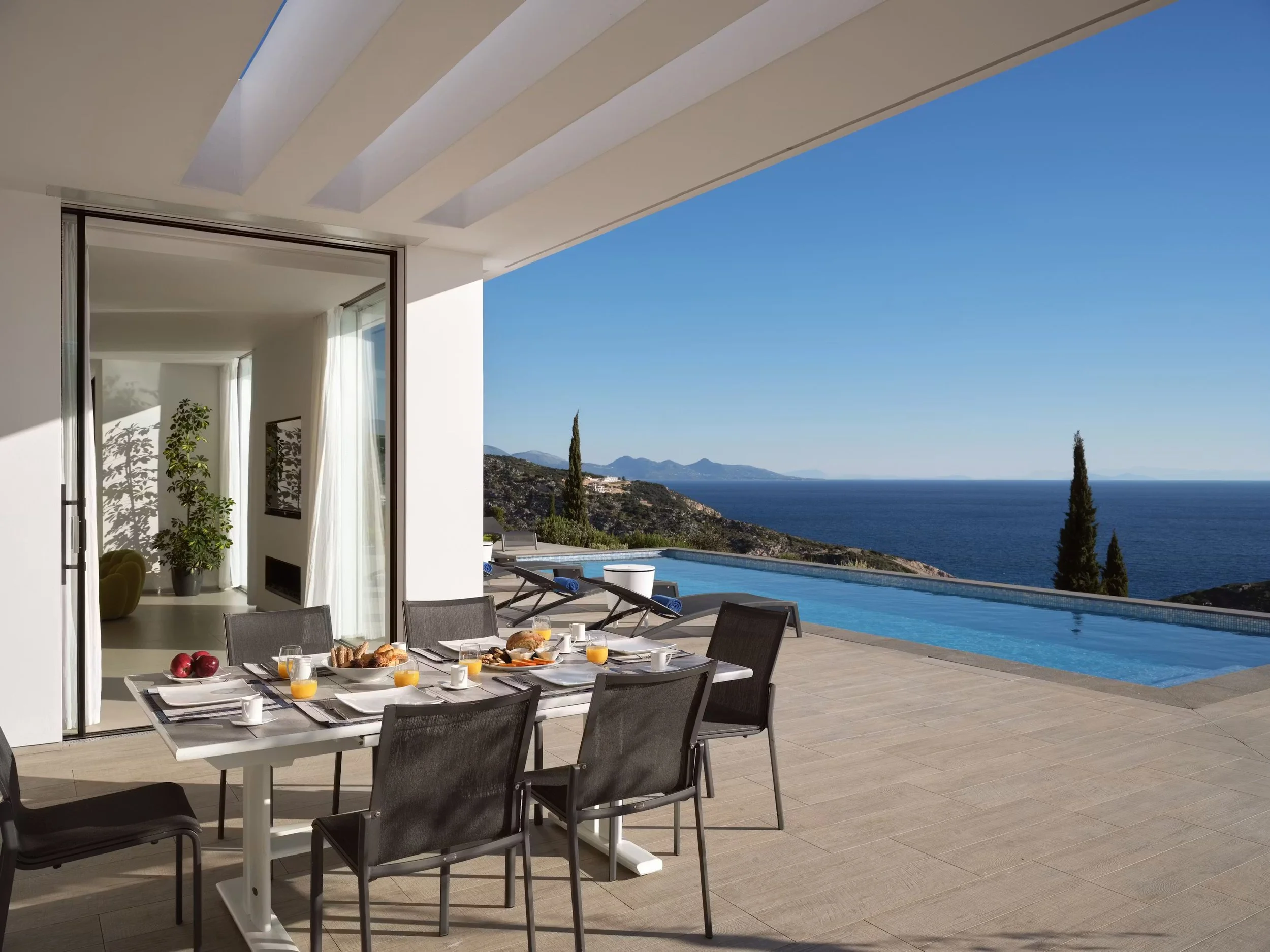 An outdoor patio with a dining table set for breakfast, overlooking a scenic view of the ocean, mountains, and a clear blue sky.