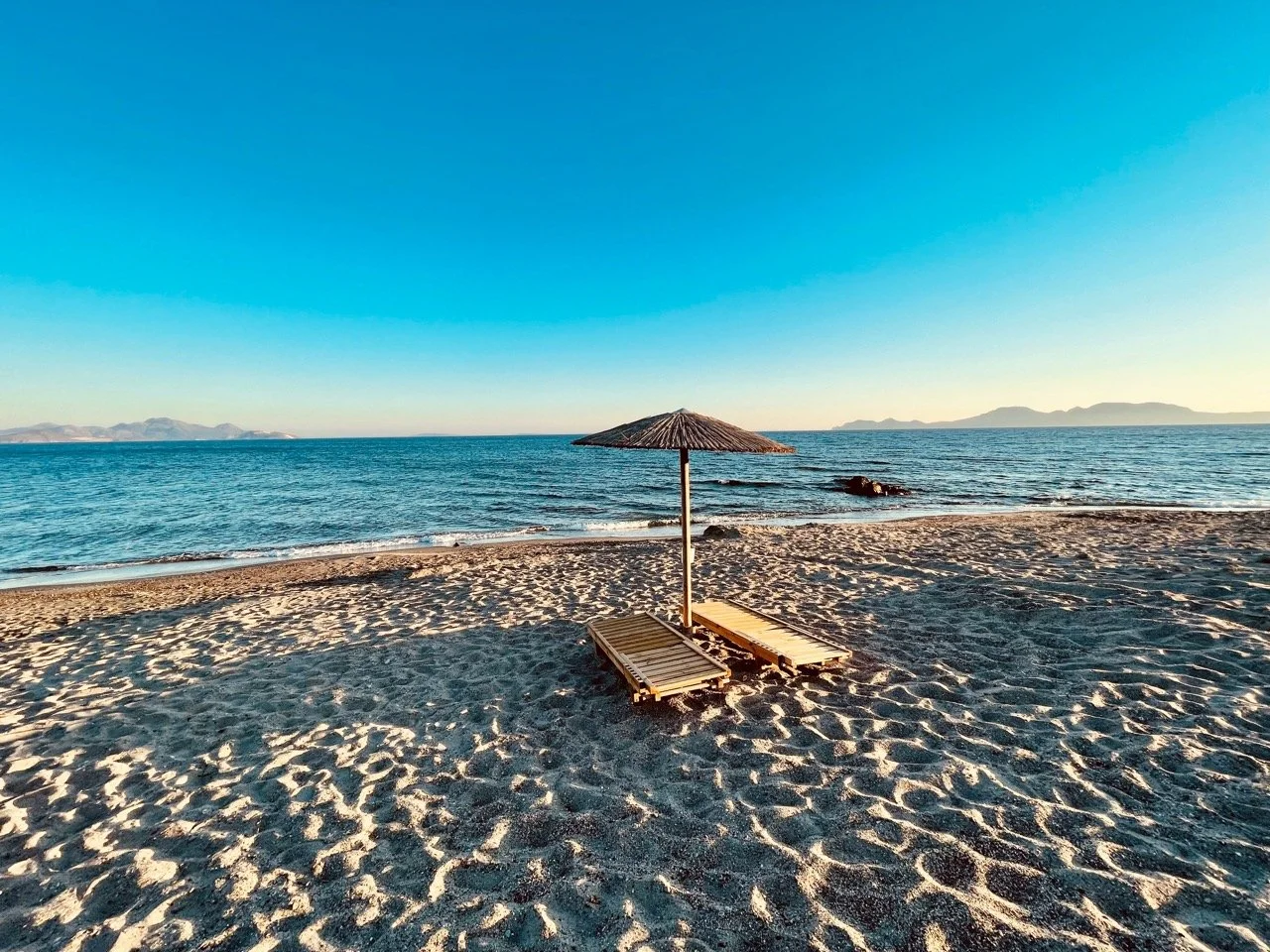 Empty beach with two wooden lounge chairs beneath a thatched umbrella, ocean in the background, clear blue sky with distant mountains.