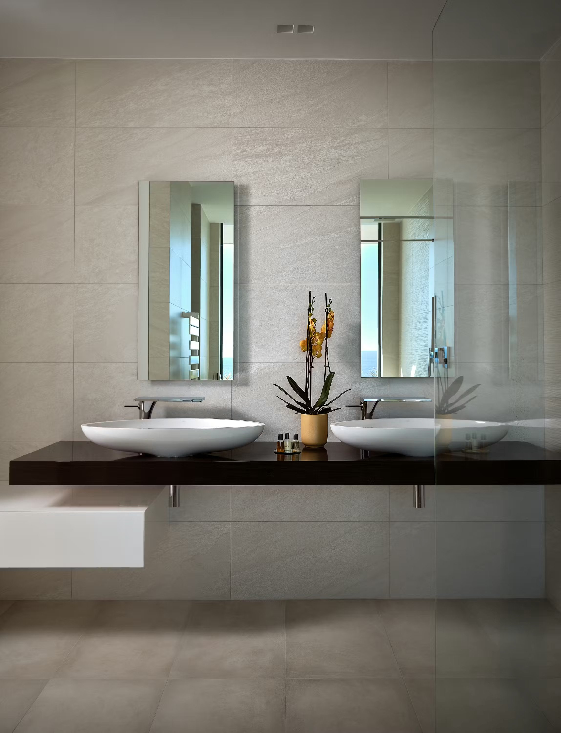 Modern bathroom with a double vanity, two oval vessel sinks, and large mirrors, decorated with a yellow orchid plant and toiletries, with natural light coming through a window.