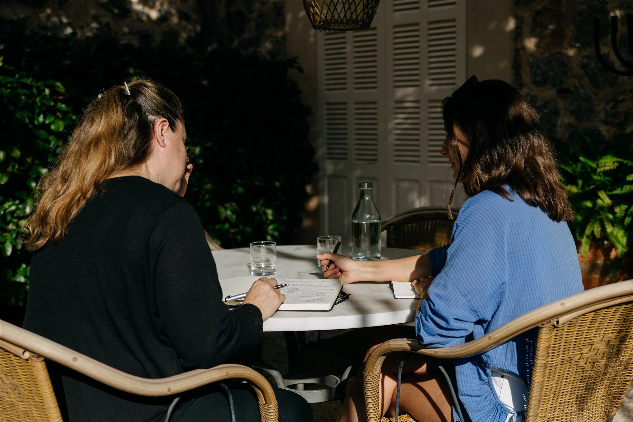 Two women sitting at a table outdoors, engaged in conversation, with notebooks and a pen, a glass of water, and a bottle, in a dimly lit setting surrounded by greenery.
