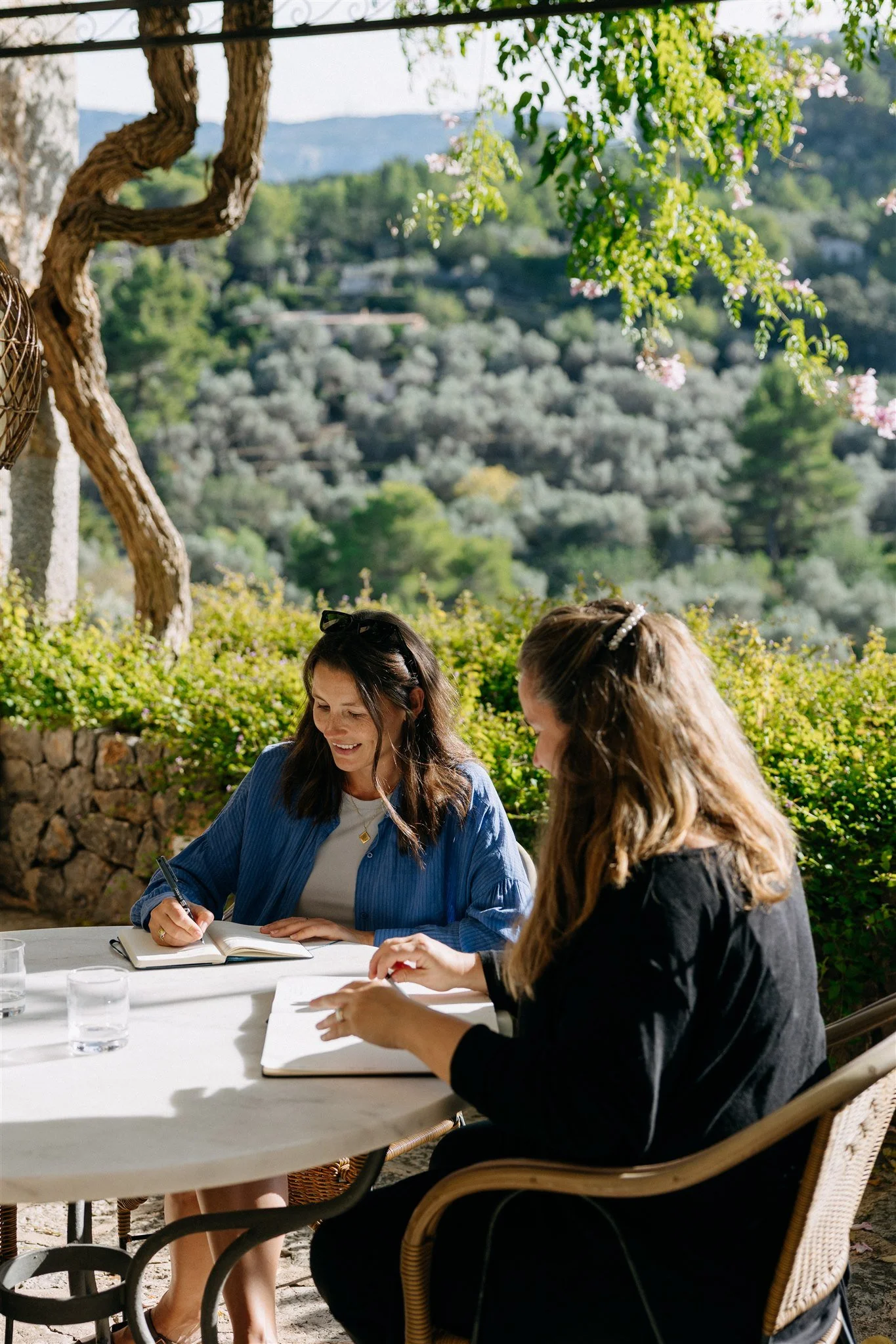 Two women sitting at a white outdoor table, smiling and writing in notebooks, with a scenic background of trees and hills