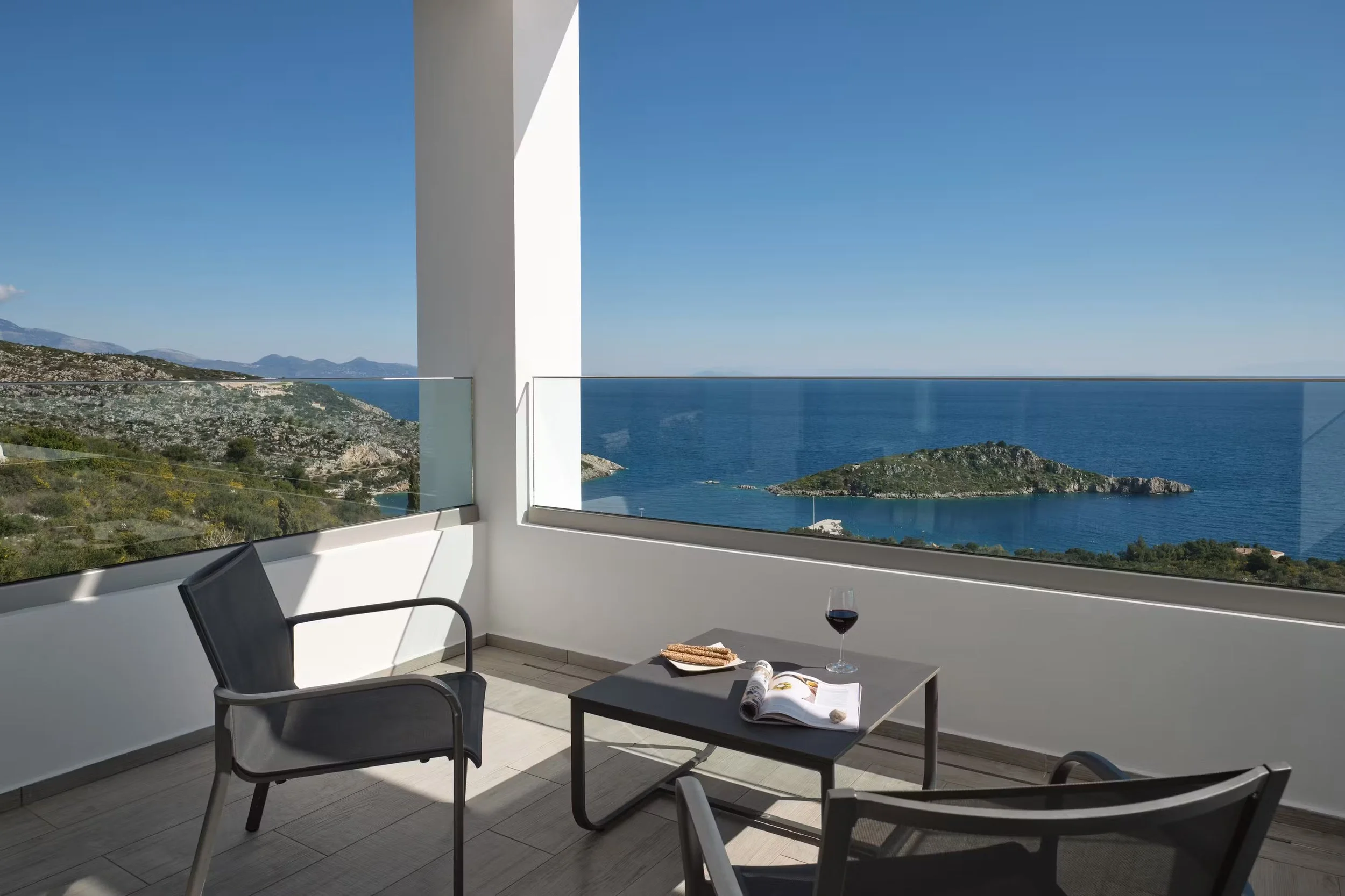 Balcony with a view of the ocean, mountains, and blue sky, furnished with a black chair, a small table with a glass of red wine, an open magazine, and some breadsticks, framed by white walls and glass railings.