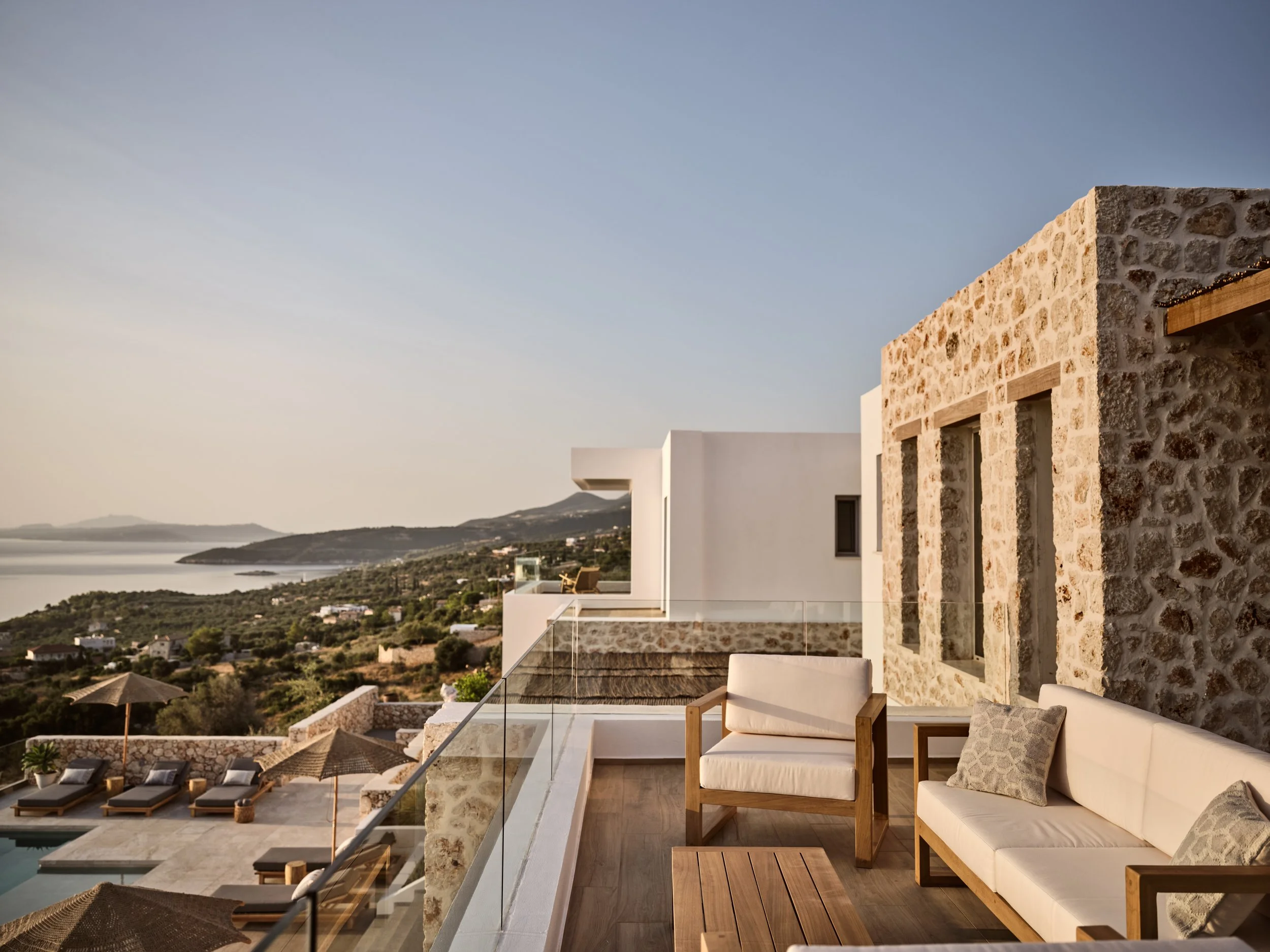An outdoor seating area on a terrace with a white sofa, wooden chairs, and throw pillows, overlooking a landscape with water and hills under a clear sky.