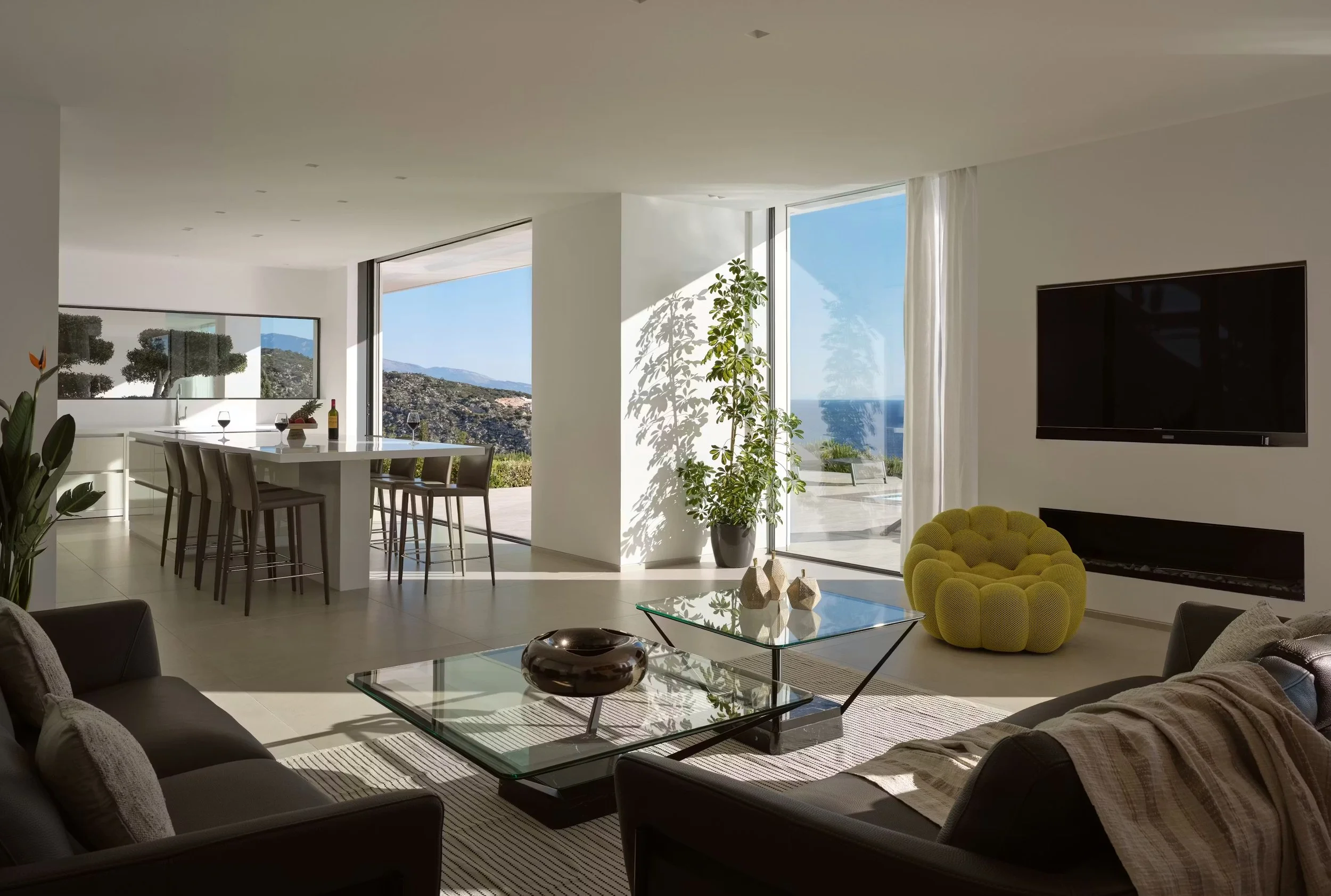 Modern living room with large windows showing a view of mountains and the ocean, furnished with gray couches, a glass coffee table, a yellow textured armchair, a large flat-screen TV, and decorative items.