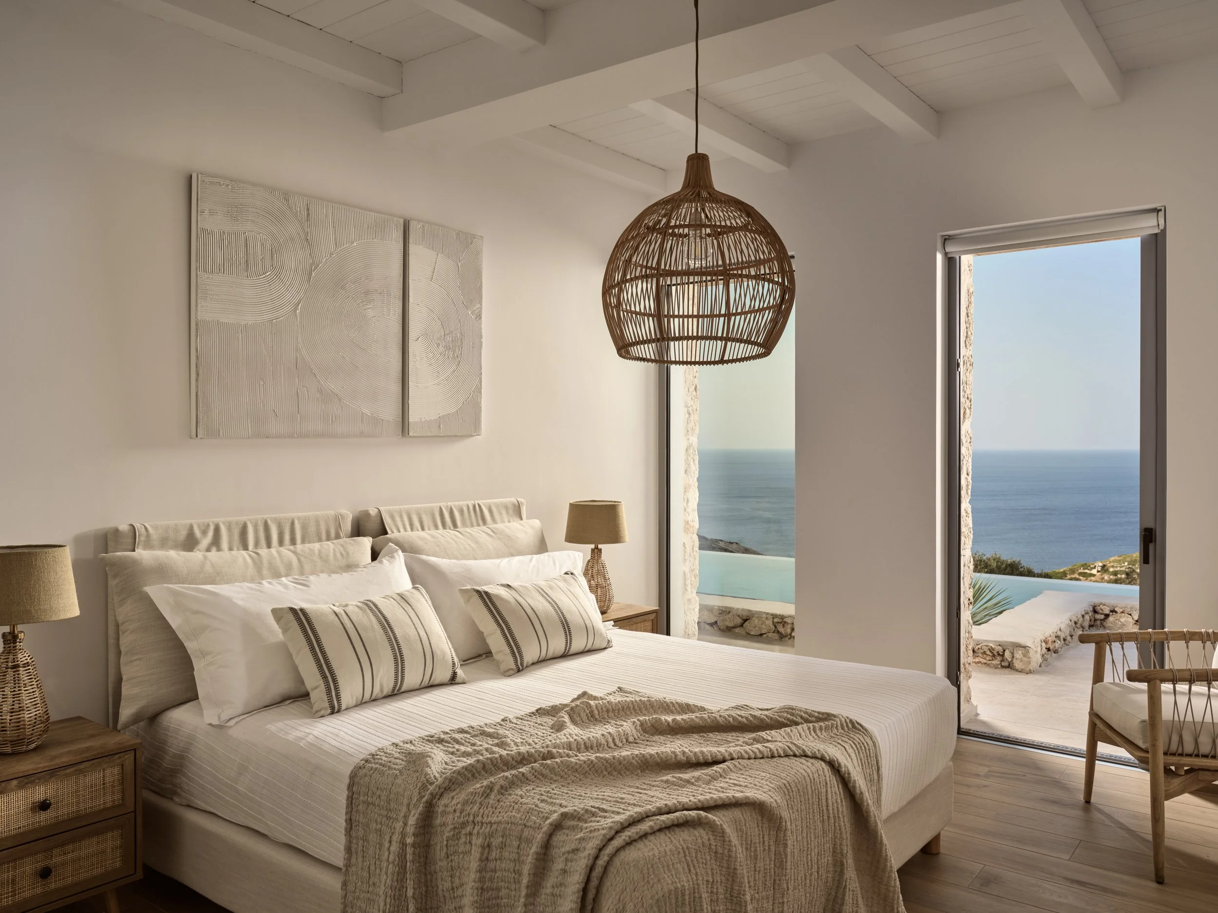 A cozy bedroom with a large white bed, striped pillows, wooden nightstand, lamps, and a woven pendant light hanging from the ceiling. The room has white walls and a view of the ocean through open windows.
