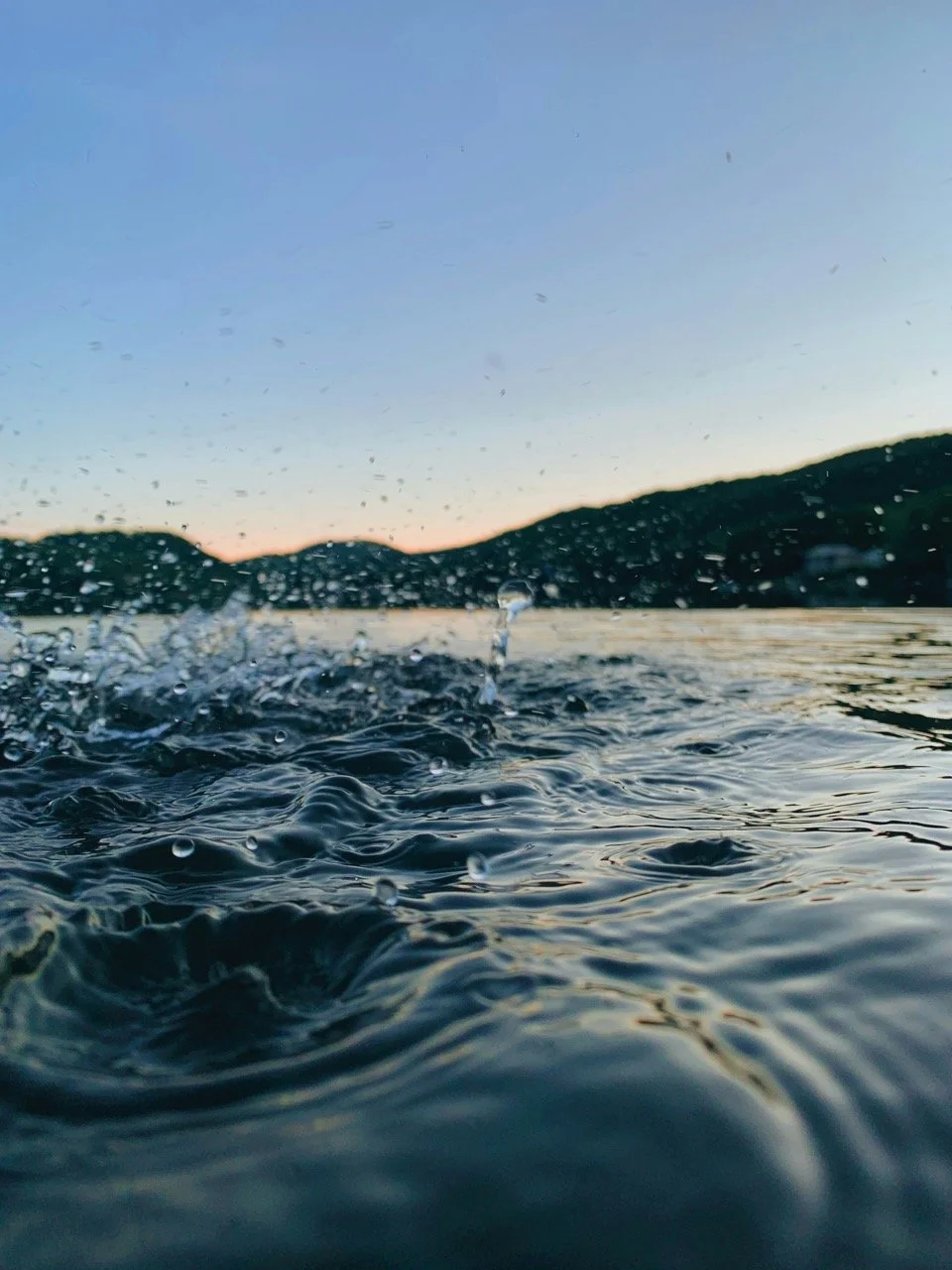 Close-up view of water with ripples and splashes, distant hills, and a clear sky during sunset or sunrise.