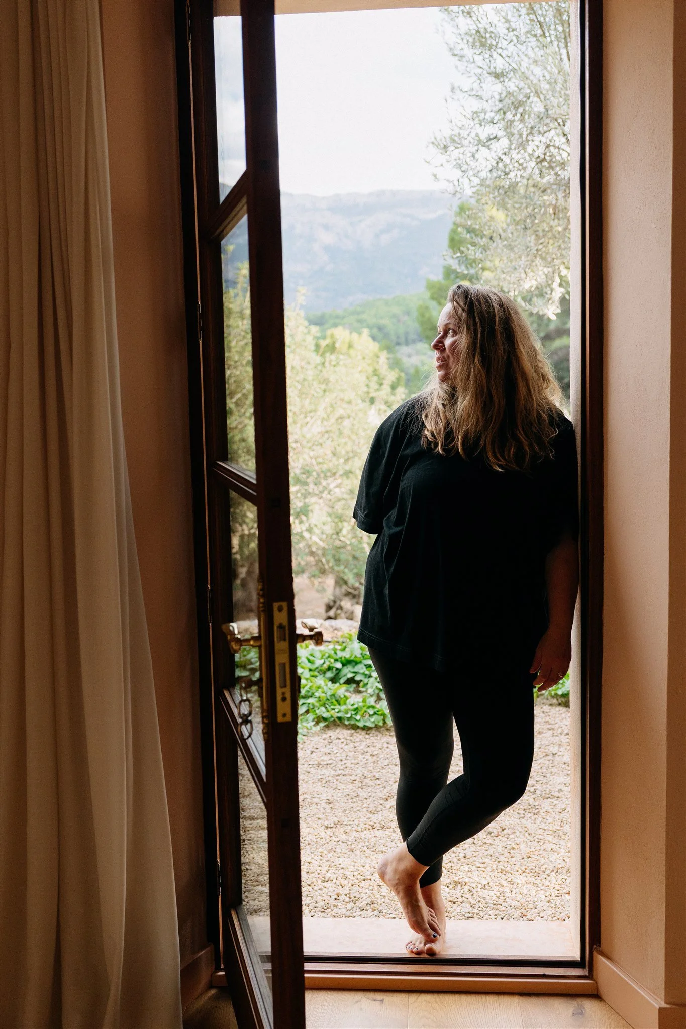 Woman standing barefoot at an open door, looking outside at trees and mountains.
