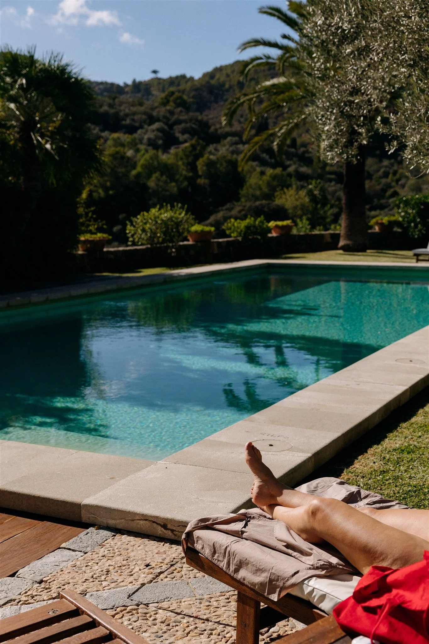 Person relaxing on a lounge chair beside a swimming pool with trees and hills in the background.