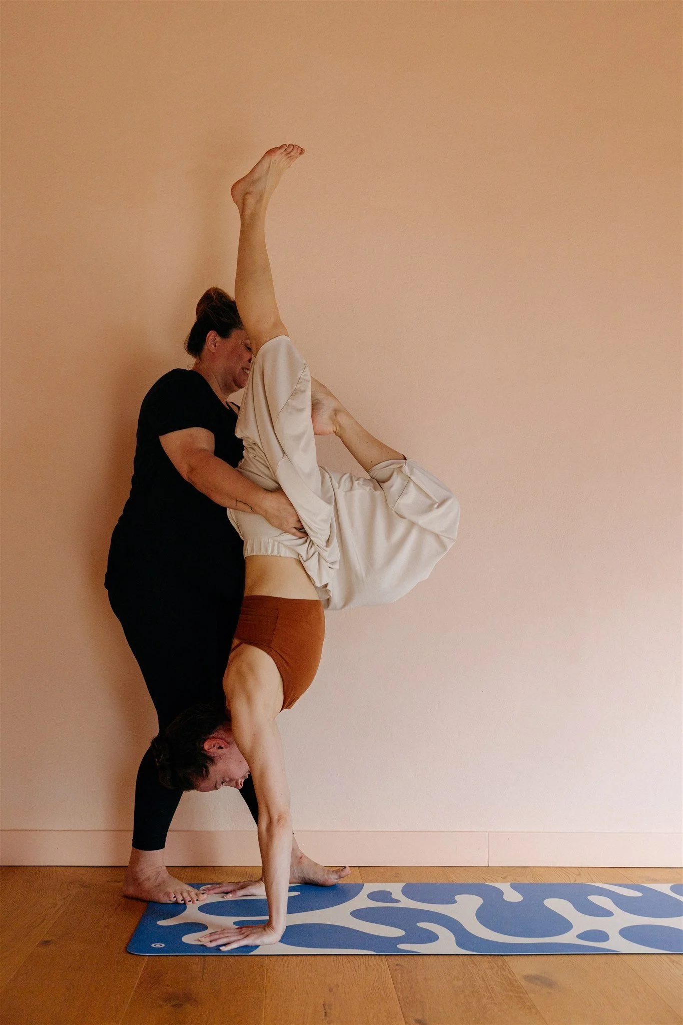 A woman practicing yoga upside down in a handstand with a partner supporting her, against a peach-colored wall.