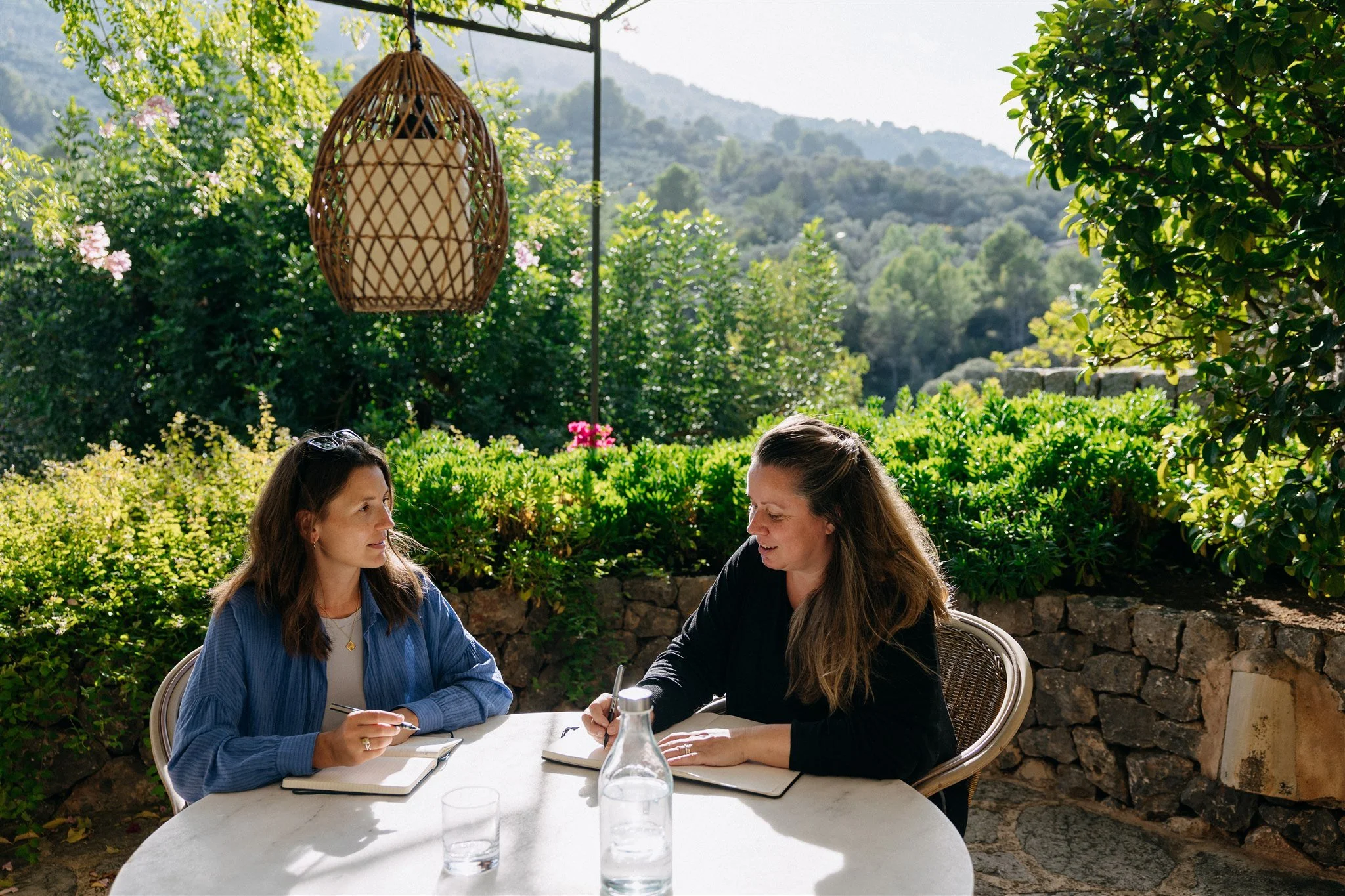 Two women sitting at a round outdoor table having a conversation, with notebooks and a glass water pitcher on the table, in a lush green garden with mountains in the background during daytime.