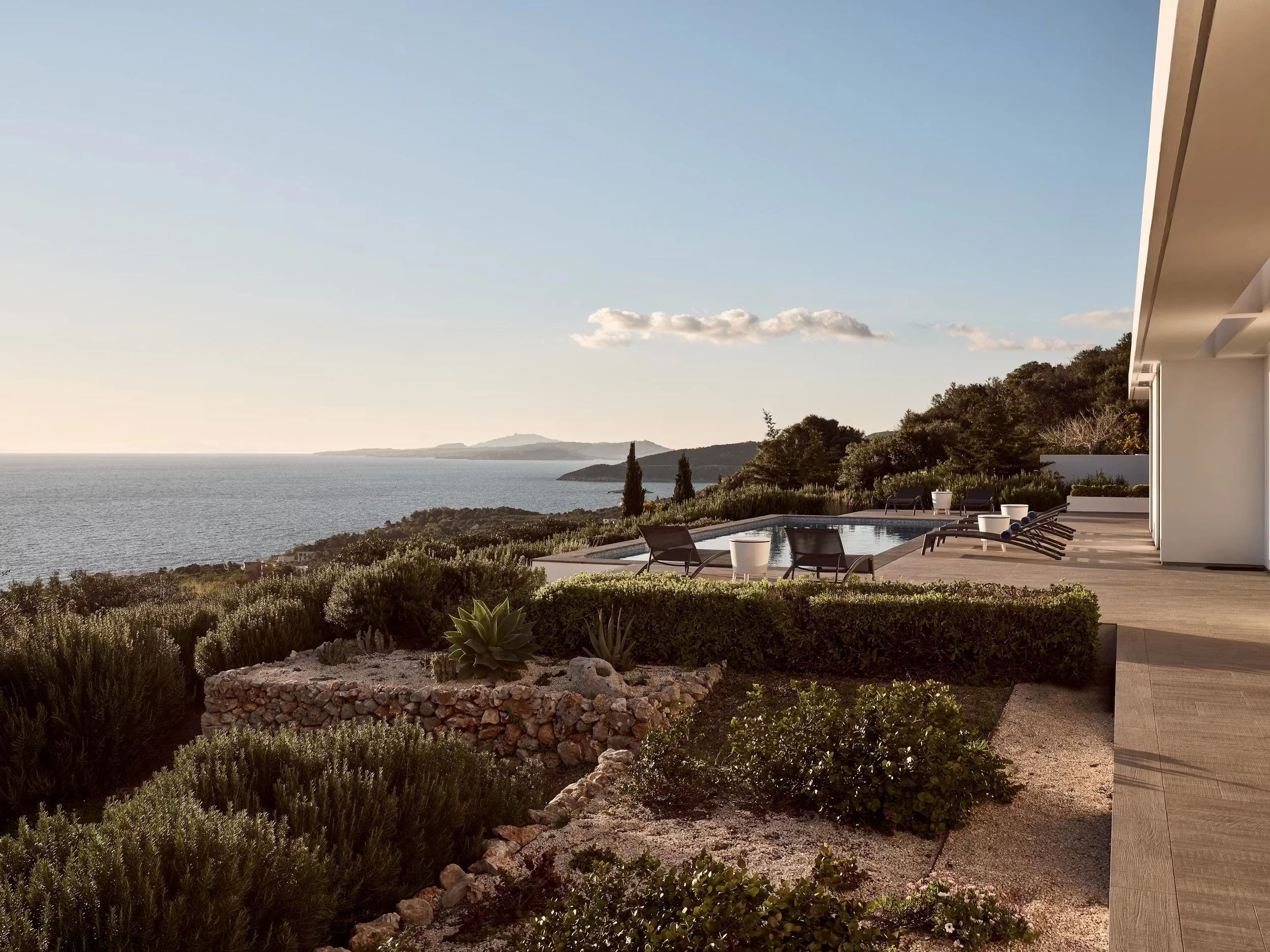 An outdoor patio area with lounge chairs, a small swimming pool, lush greenery, and a view of a large body of water with distant hills, under a clear sky with a few clouds.