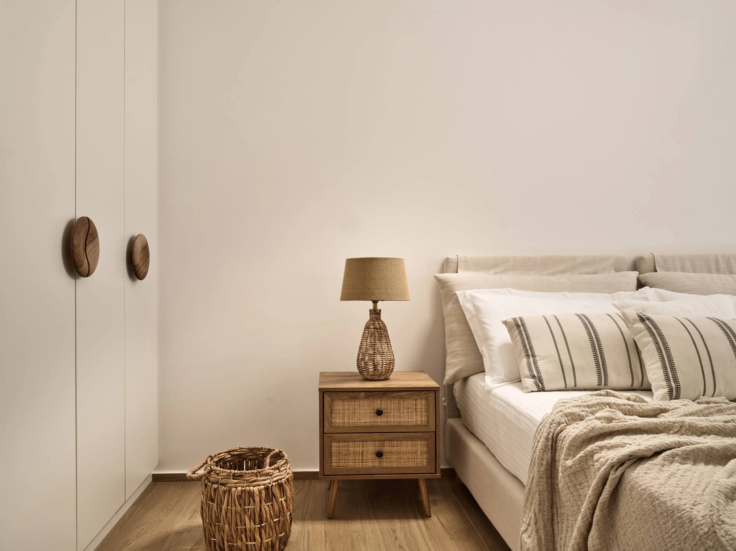 A bedroom with a beige bed, striped bedding, a wooden nightstand with rattan drawers, a wicker table lamp, a woven basket, and a white wardrobe with wooden oval handles.