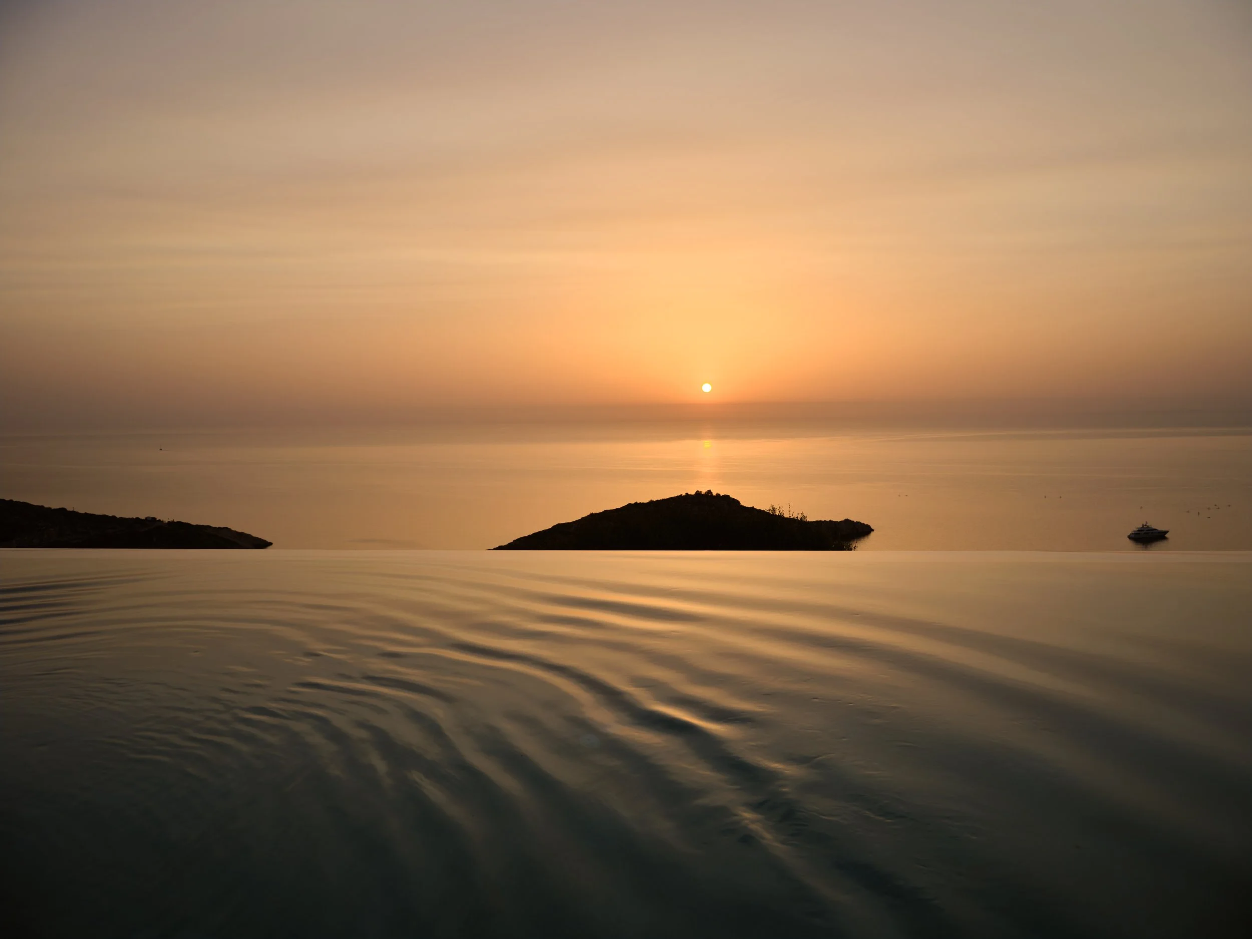 A sunset over the ocean with the sun near the horizon, a small boat on the water, and a silhouette of a small island or landmass in the distance.