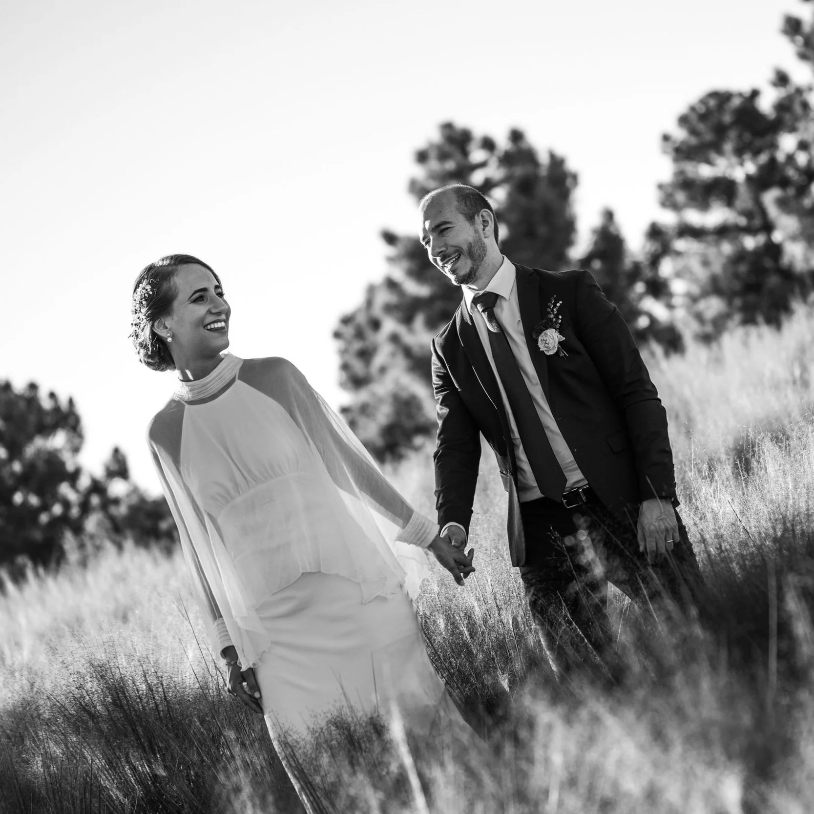 Pareja recién casada caminando en un campo, sosteniéndose de la mano y sonriendo, en una foto en blanco y negro. Bodas y eventos. Madrid