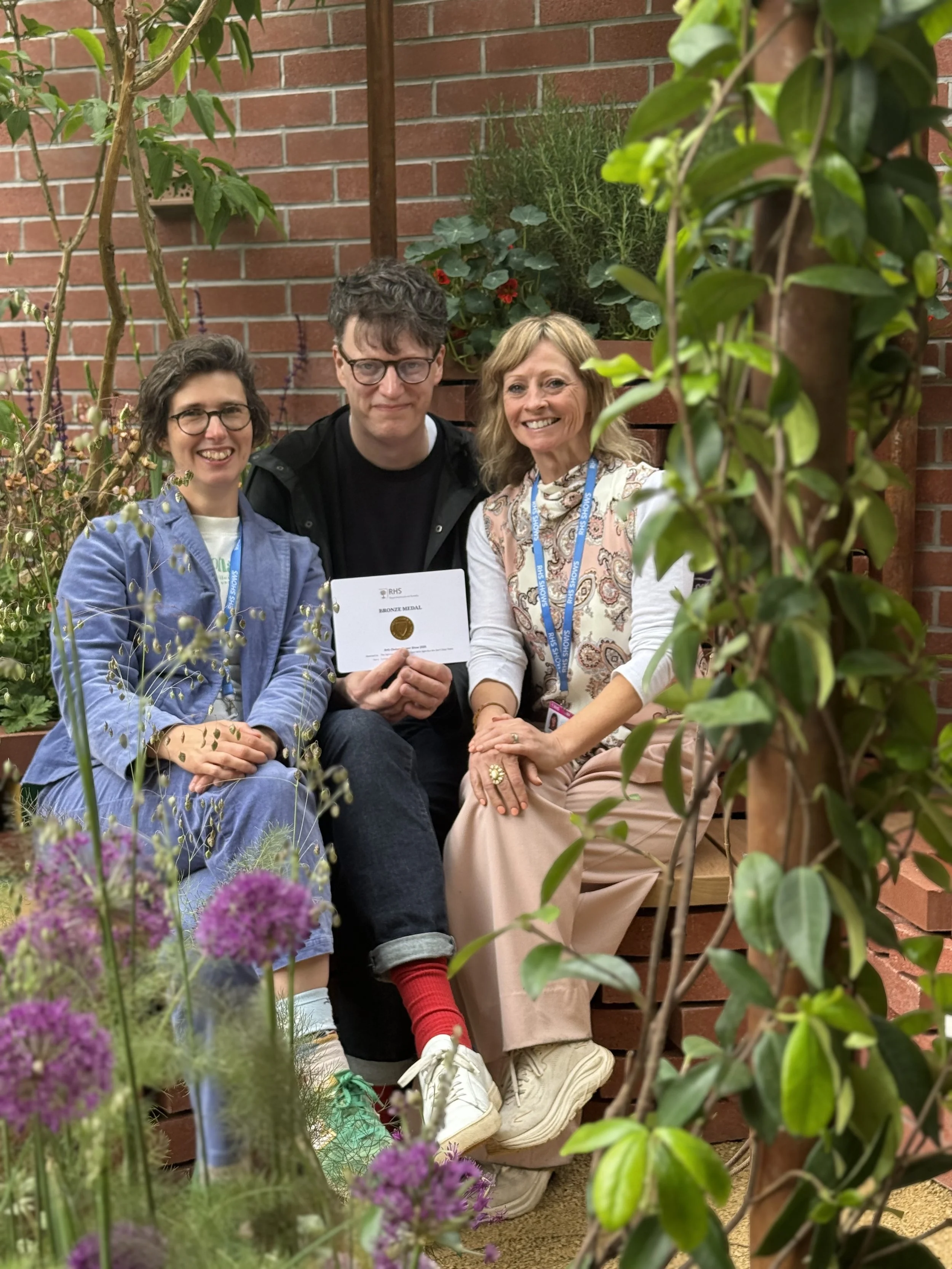 Three people sitting closely together outdoors, surrounded by green plants and flowers, holding a certificate or medal, smiling at the camera.