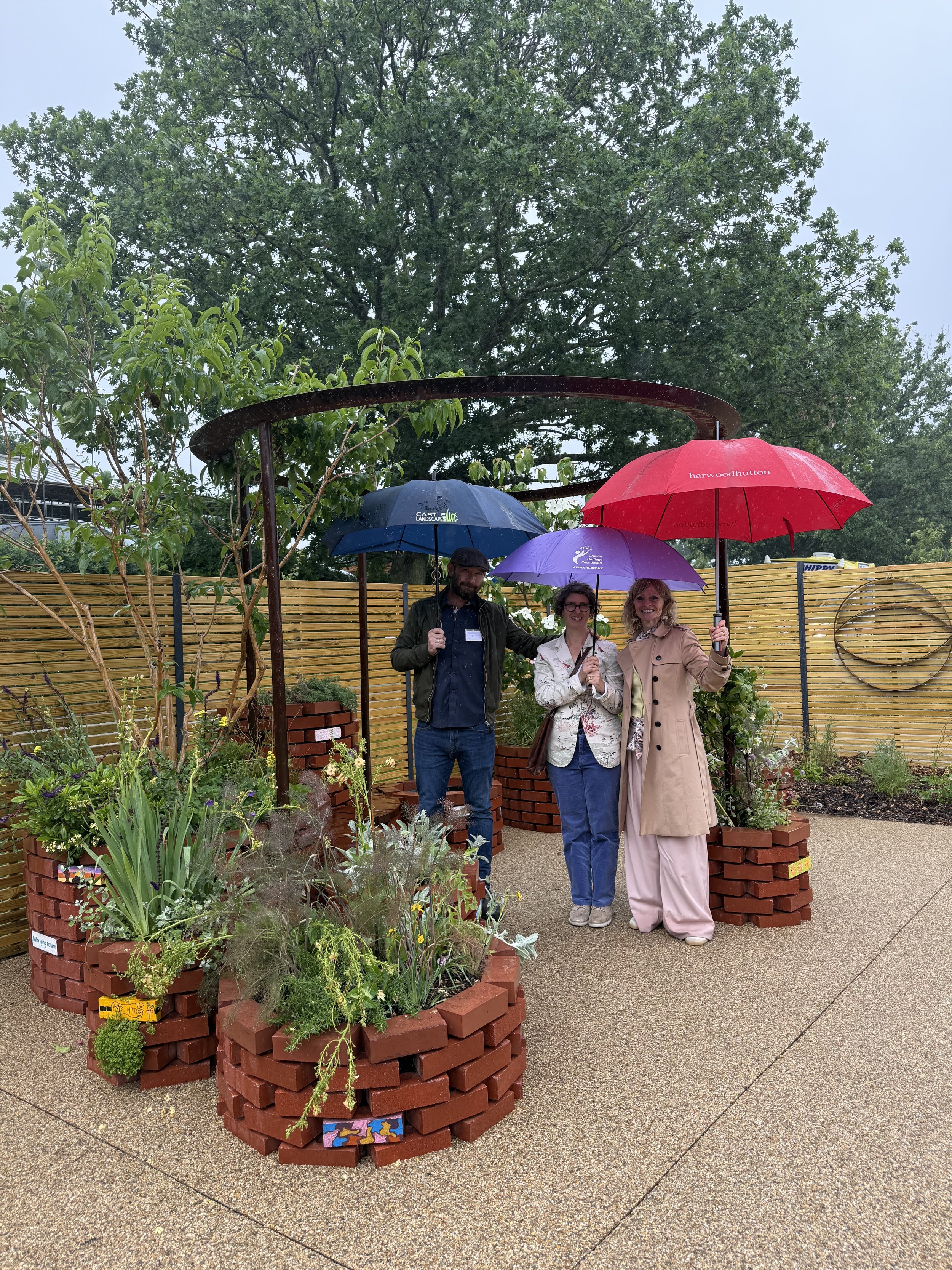 Three people holding umbrellas stand among brick planter beds filled with plants and flowers, with a wooden fence behind them, on a rainy day in a garden or outdoor space.