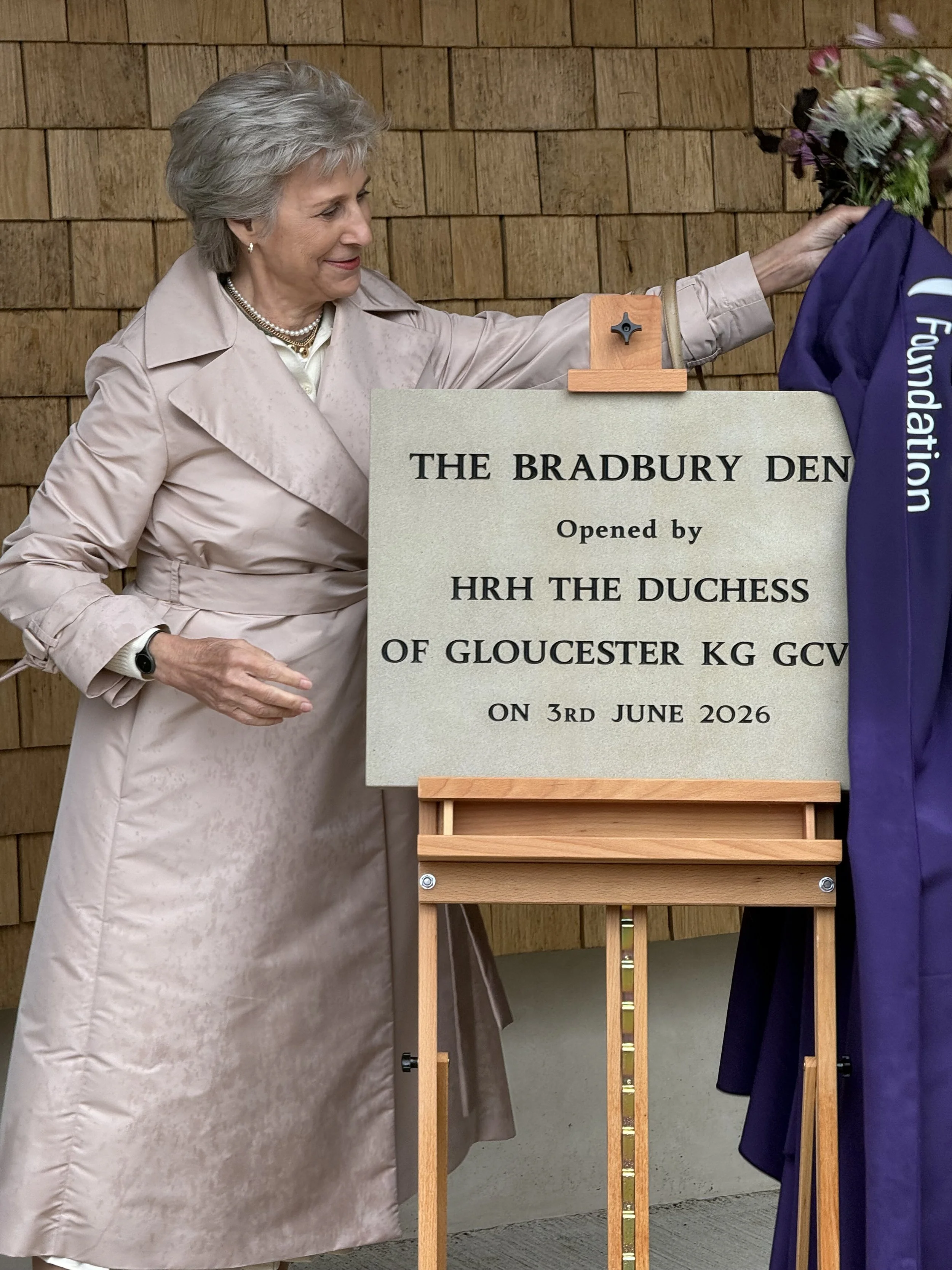 Queen Elizabeth II participating in the opening of The Bradbury Den, holding a bouquet of flowers while standing near a sign about the event on June 3rd, 2026.