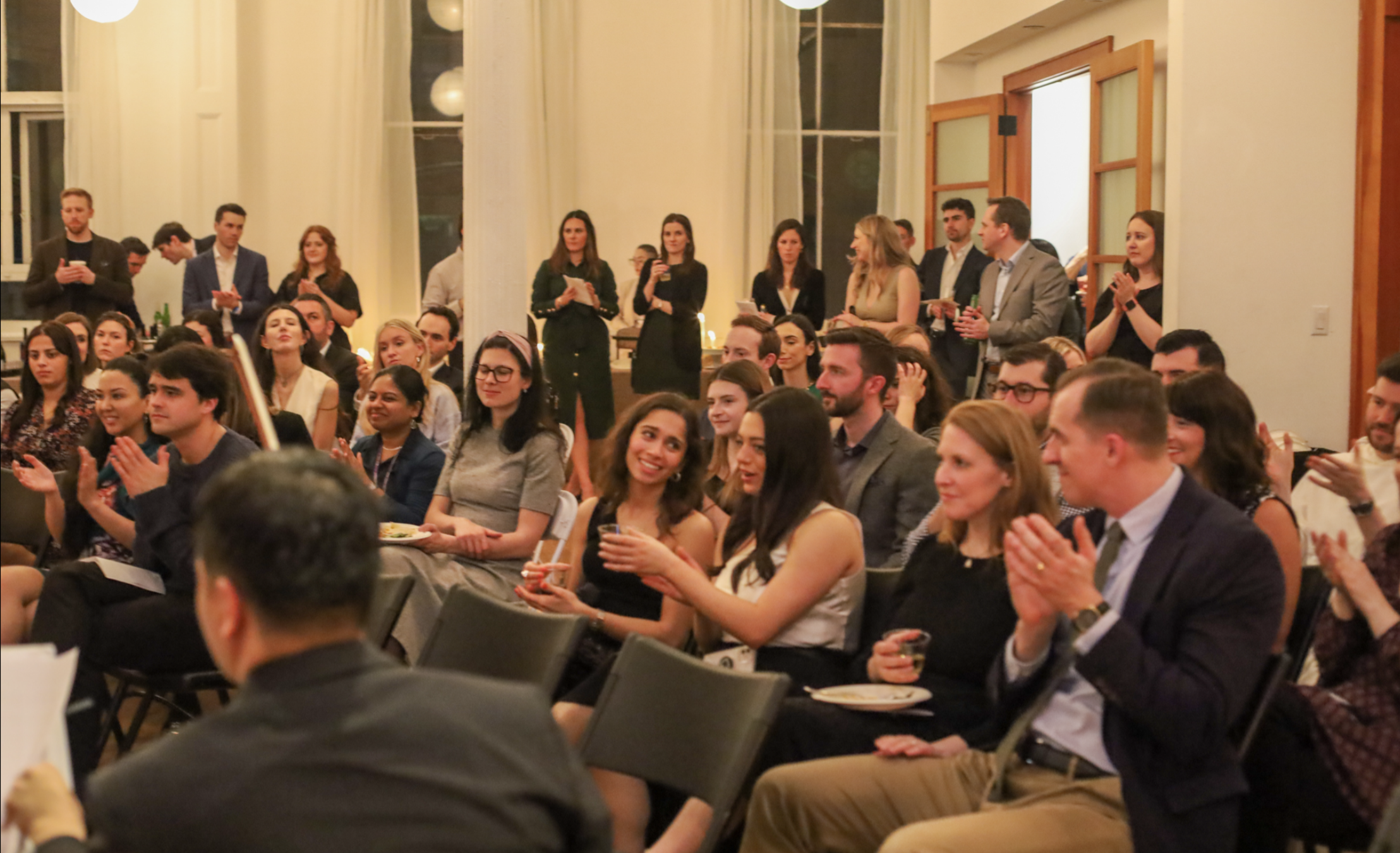 A large group of people attending a formal event in a spacious, well-lit room with high ceilings, large windows, and elegant curtains. Some are seated, smiling, and clapping, while others stand along the walls, some holding drinks or taking photos.