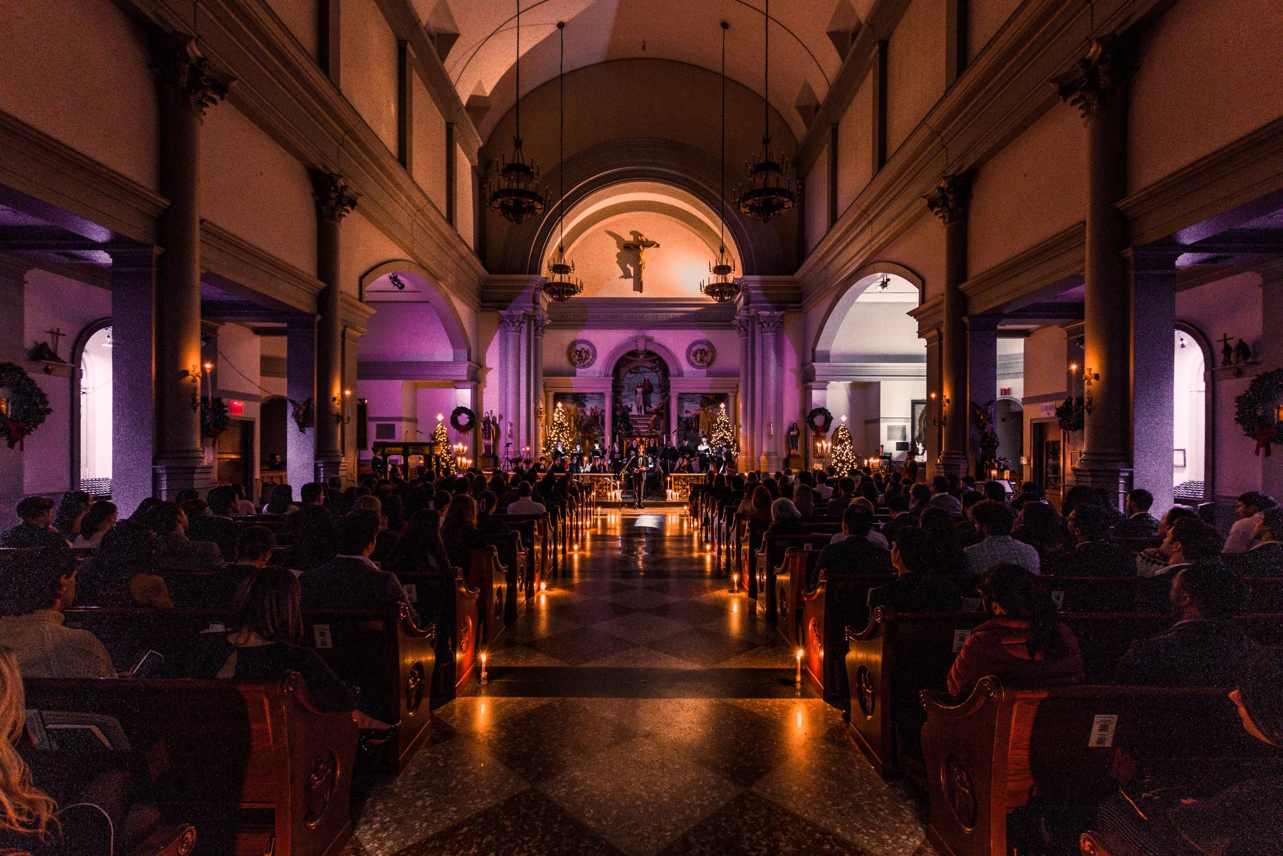 Interior of a church decorated for Christmas with Christmas trees, wreaths, and lights, during a concert or service with an audience.