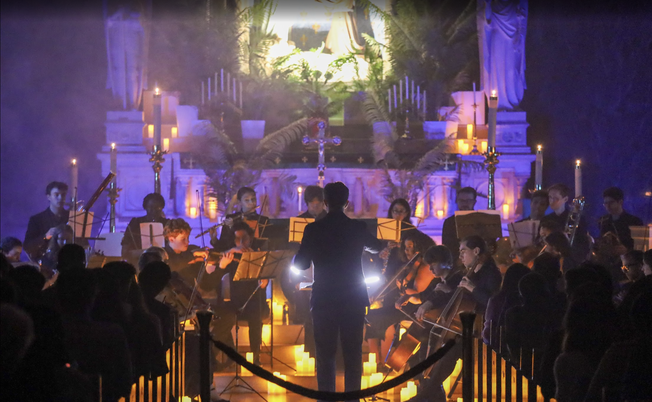 Orchestra playing music in a church with candles and religious statues on the altar behind them.