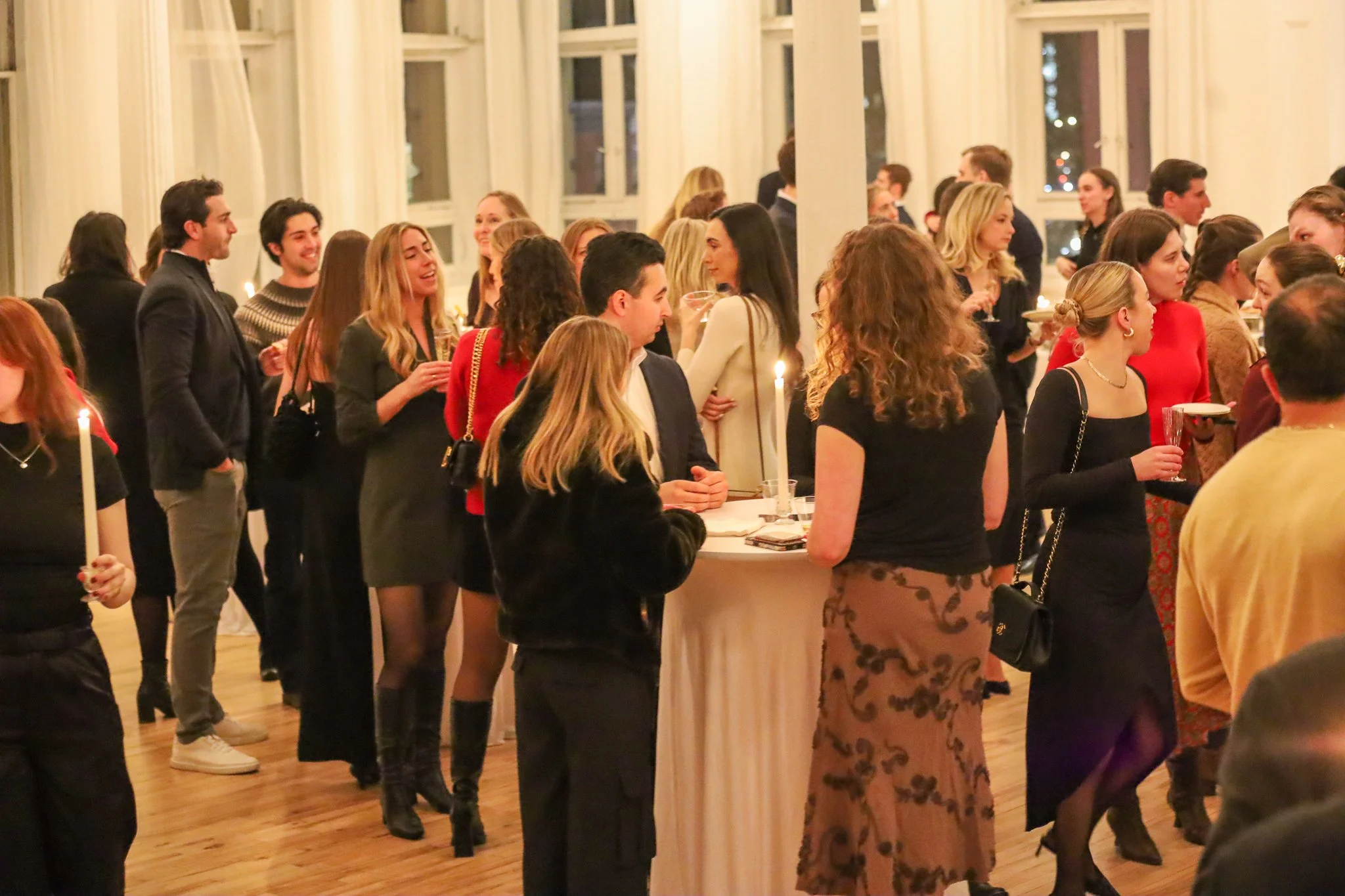 People socializing at an indoor party, standing and talking in a well-lit room with large windows and wooden floors.