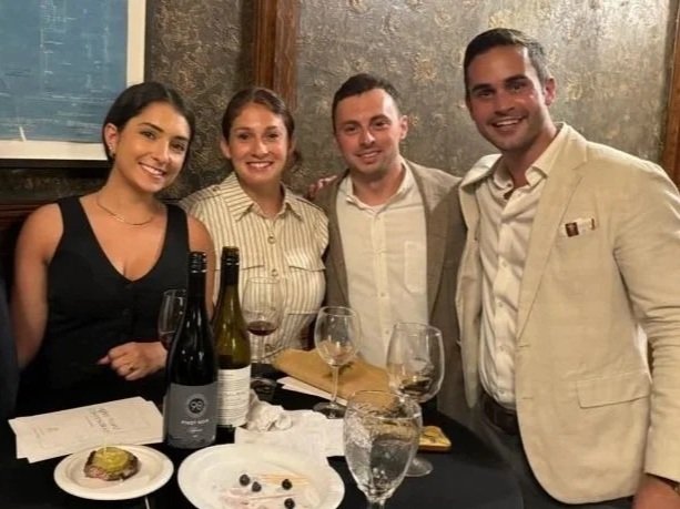 Four people smiling and posing for a photo at a dinner table with wine bottles, glasses, and dessert.