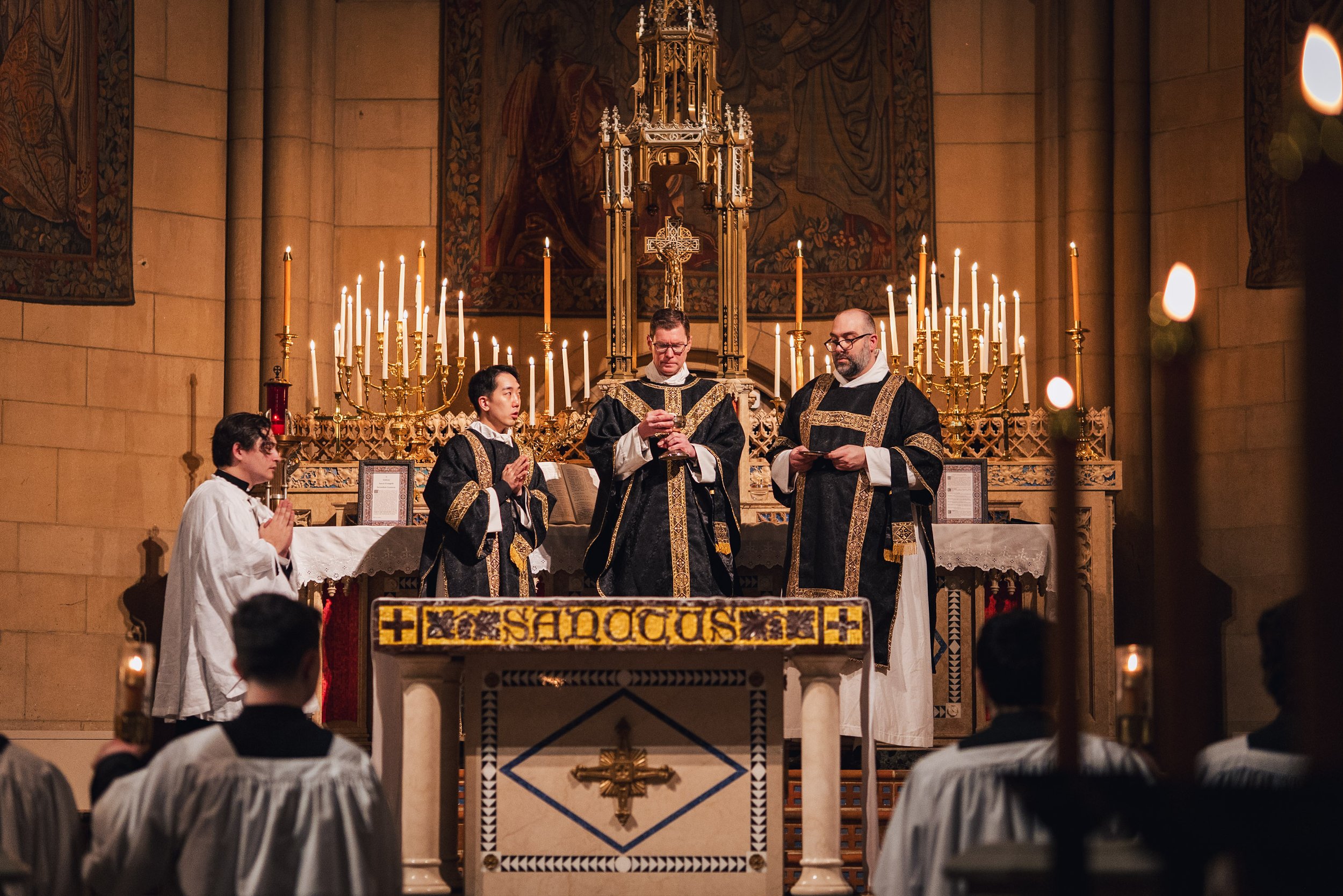 Four clergy members standing behind an altar during a religious service inside a church with candles and religious art.