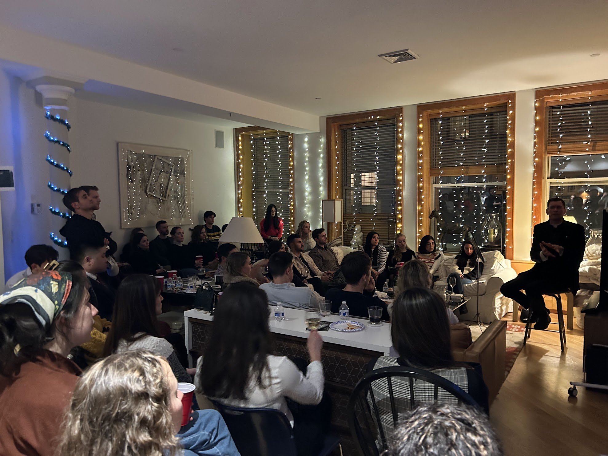 People gathering in a living room decorated with string lights, with a man speaking in front of the group. The room has large windows with blinds and wooden frames, and there is a Christmas tree visible through the window.
