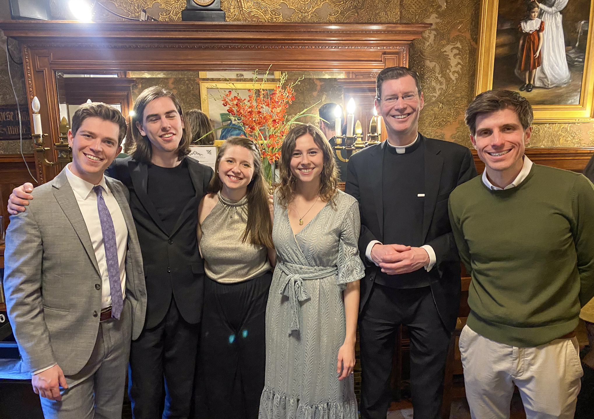 A group of six people, including three men, two women, and one person mostly out of frame, posing together in a warmly decorated room with ornate woodwork, a mirror, a flower arrangement, and paintings in the background, all smiling at the camera.