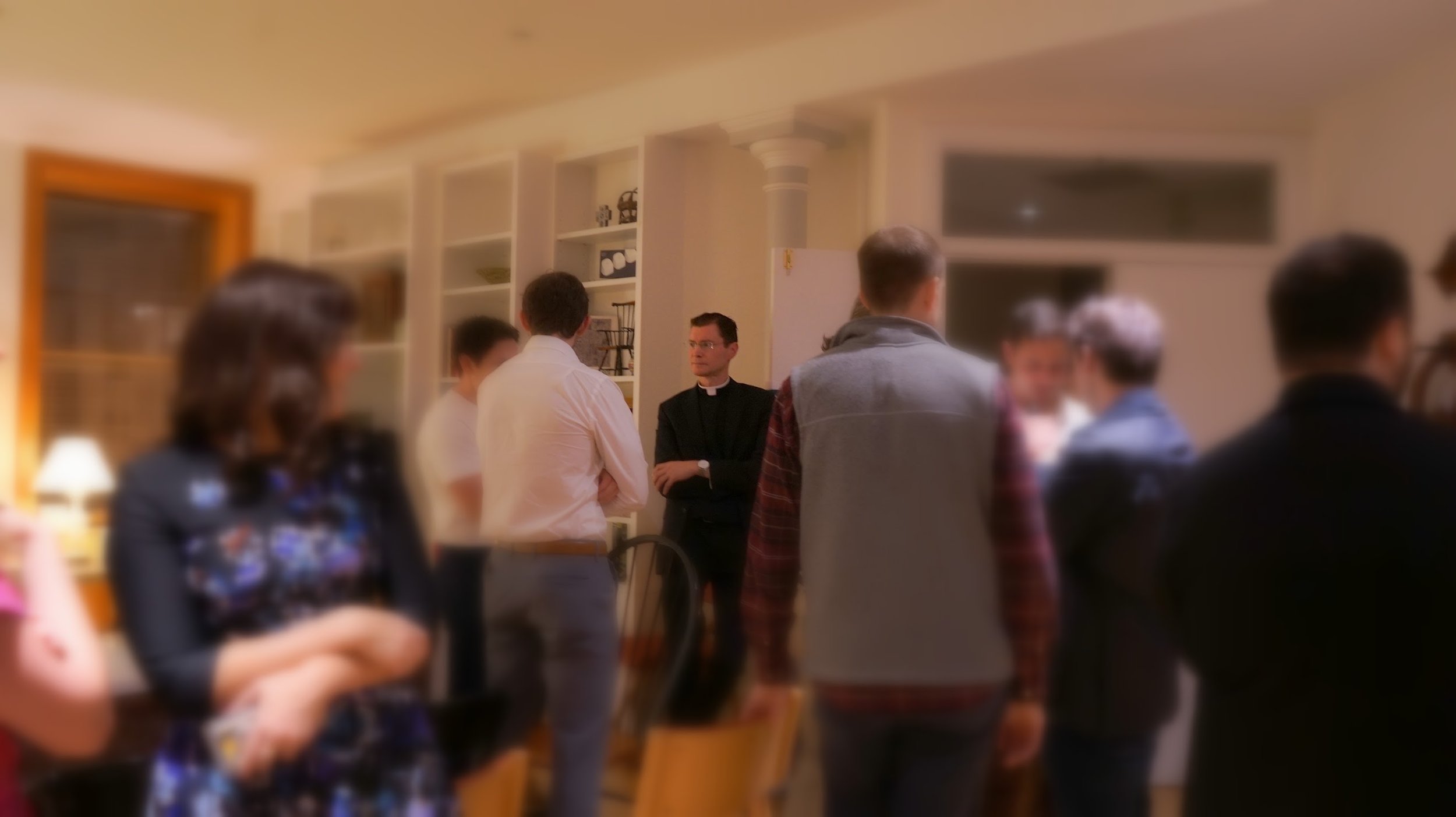 A group of people gathered indoors, engaged in conversation. A man dressed as a priest is standing in the middle, talking to others. The setting appears to be a social or community event in a room with bookshelves and warm lighting.