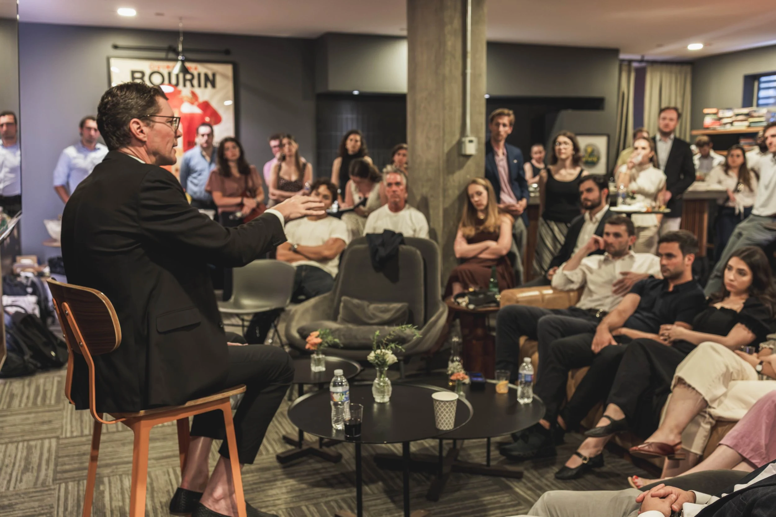 A man in a black jacket and glasses is speaking to an audience in a room with gray walls and a concrete column. The audience, seated and standing, is listening attentively. Behind the speaker, a large