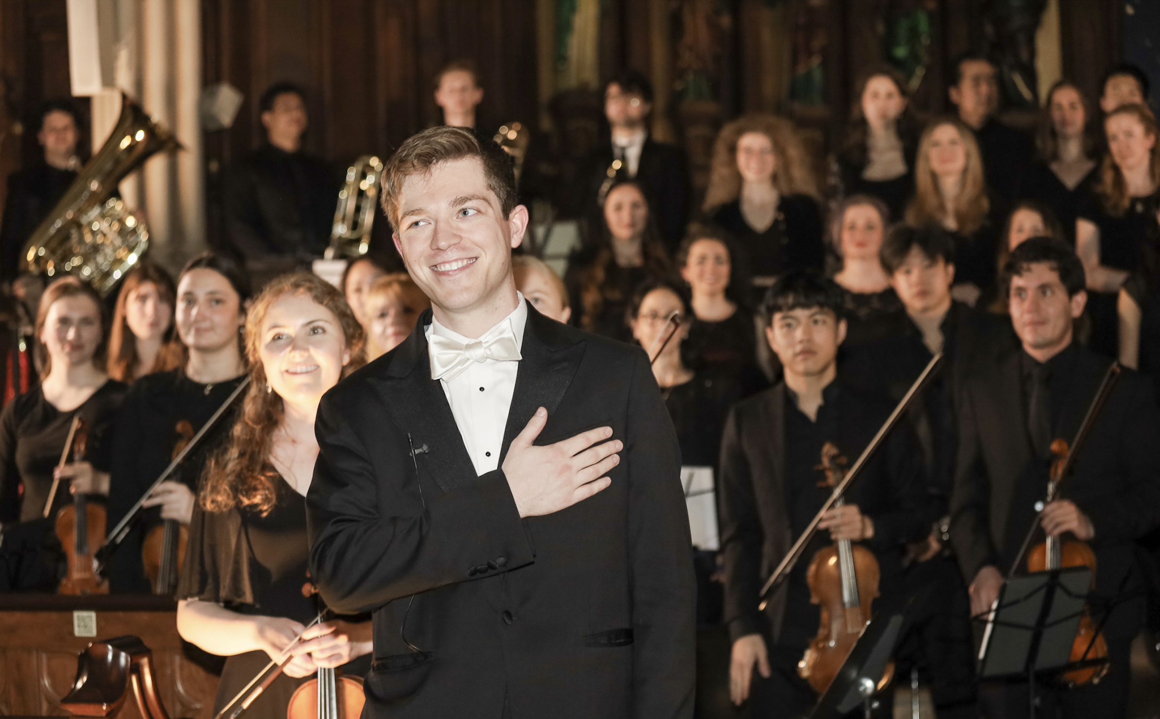 A young man in a tuxedo with a white bow tie is smiling and placing his right hand over his heart while standing in front of an orchestra, with members holding string instruments and a brass instrument visible in the background, on a stage with wood paneling.