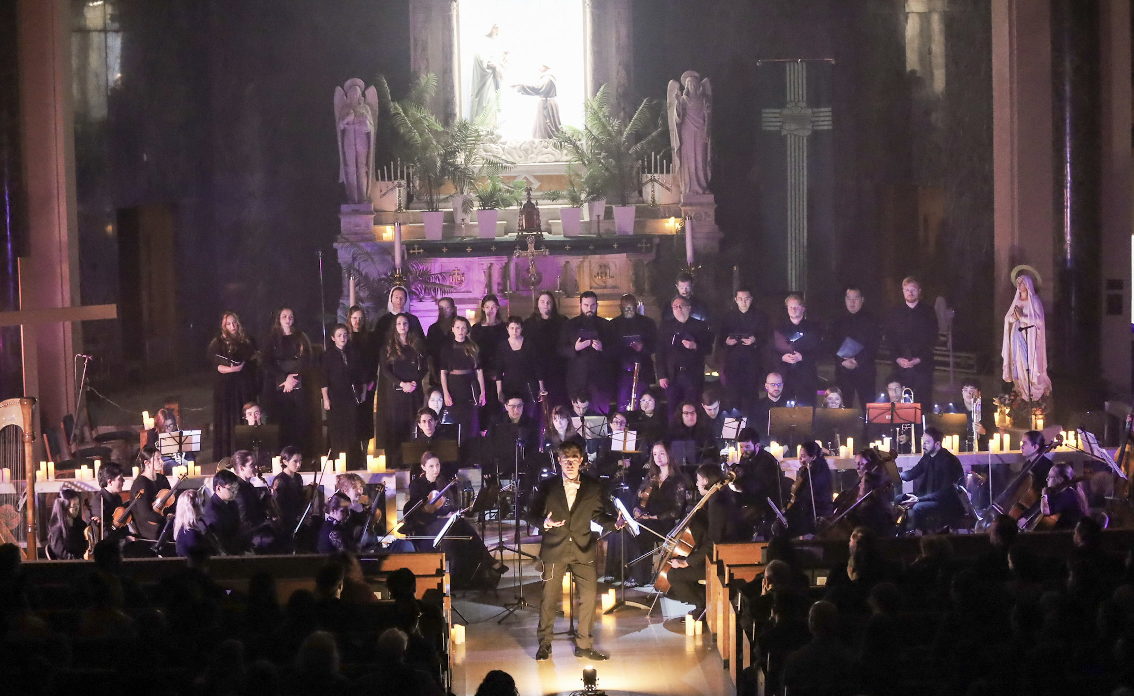 A choir and orchestra performing in a church or cathedral with an ornate altar and religious statues in the background. The conductor is standing in front, leading the performers, with candles and candles stands surrounding the stage.