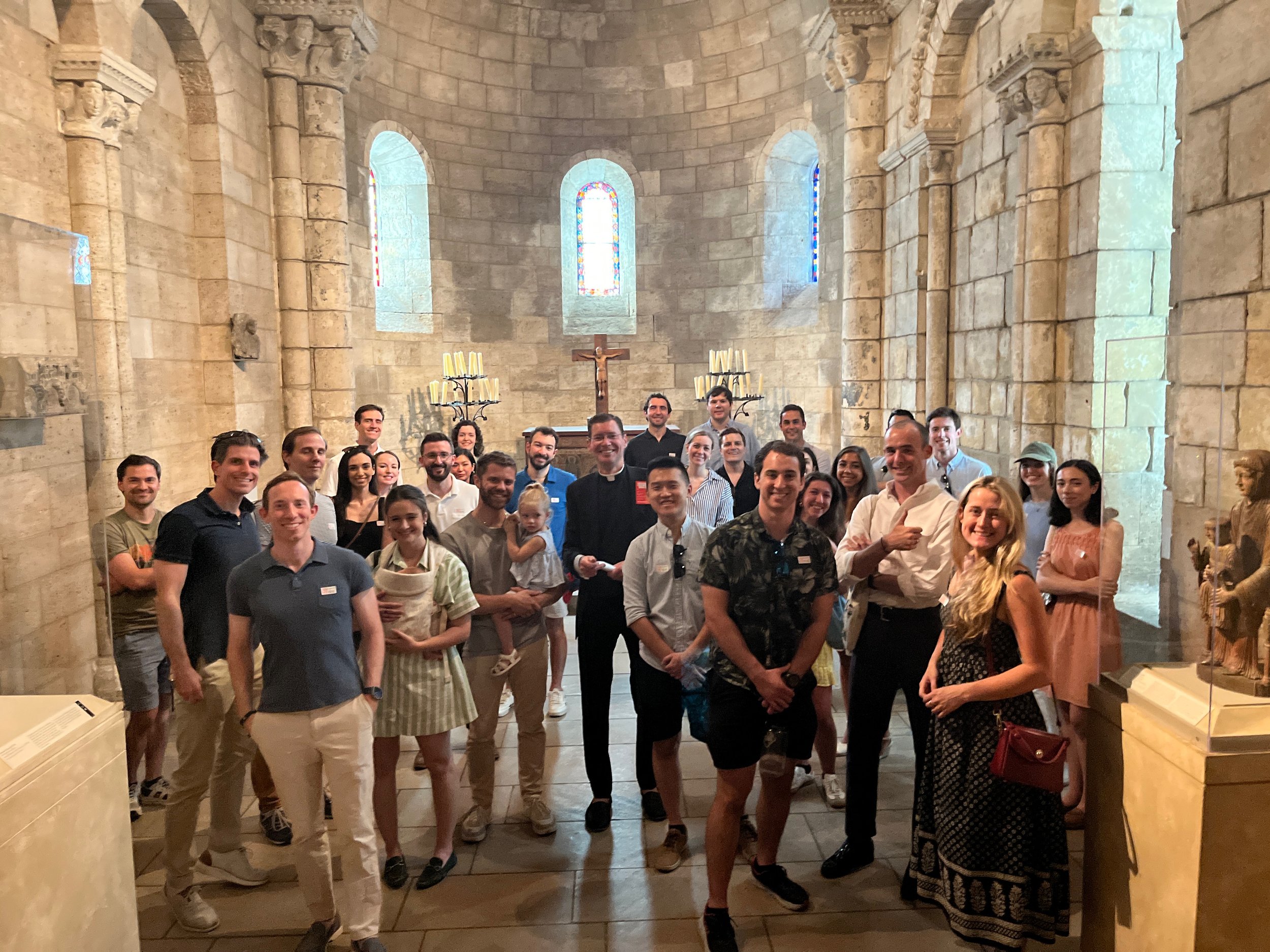A group of diverse people, including men, women, and children, standing inside a historic stone church with stained glass windows, a wooden crucifix, and lit candles, posing for a photo.