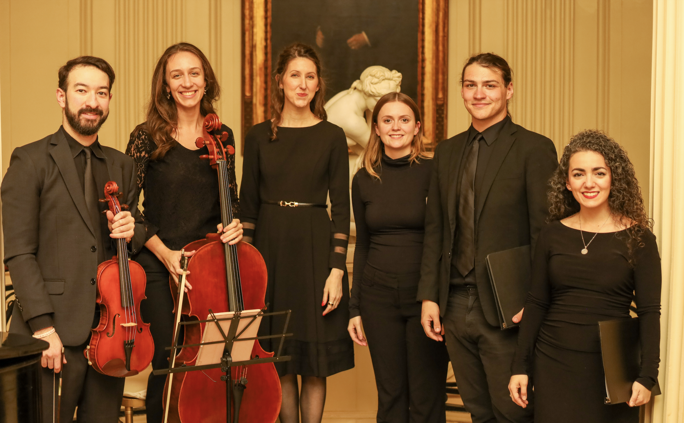Group of seven musicians, five women and two men in formal attire, standing in a room with classical decor, including a large painting and a sculpture in the background, some holding string instruments.