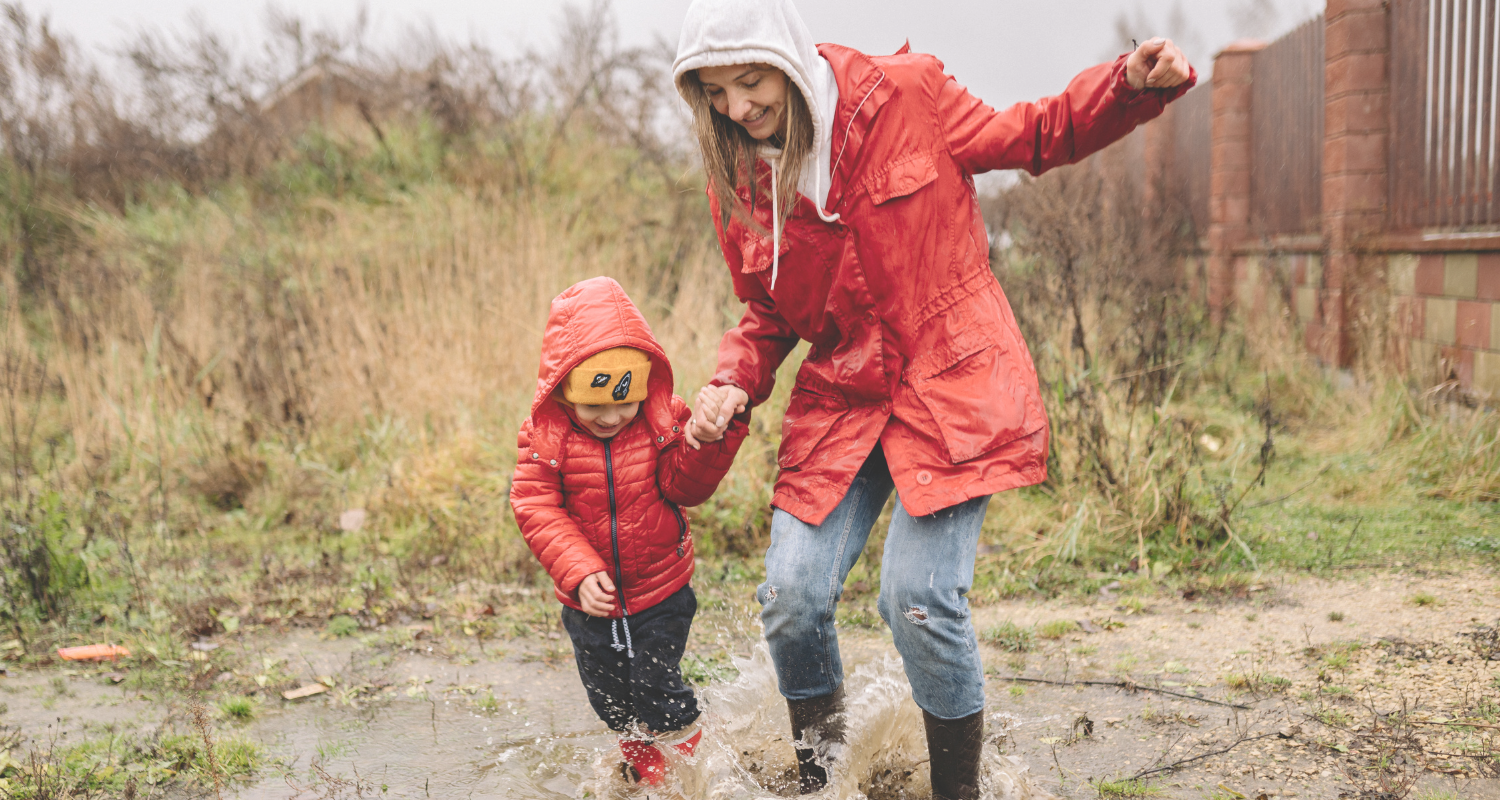 A woman and a young boy playing in a muddy puddle outdoors, holding hands and splashing water, both dressed in red jackets and boots