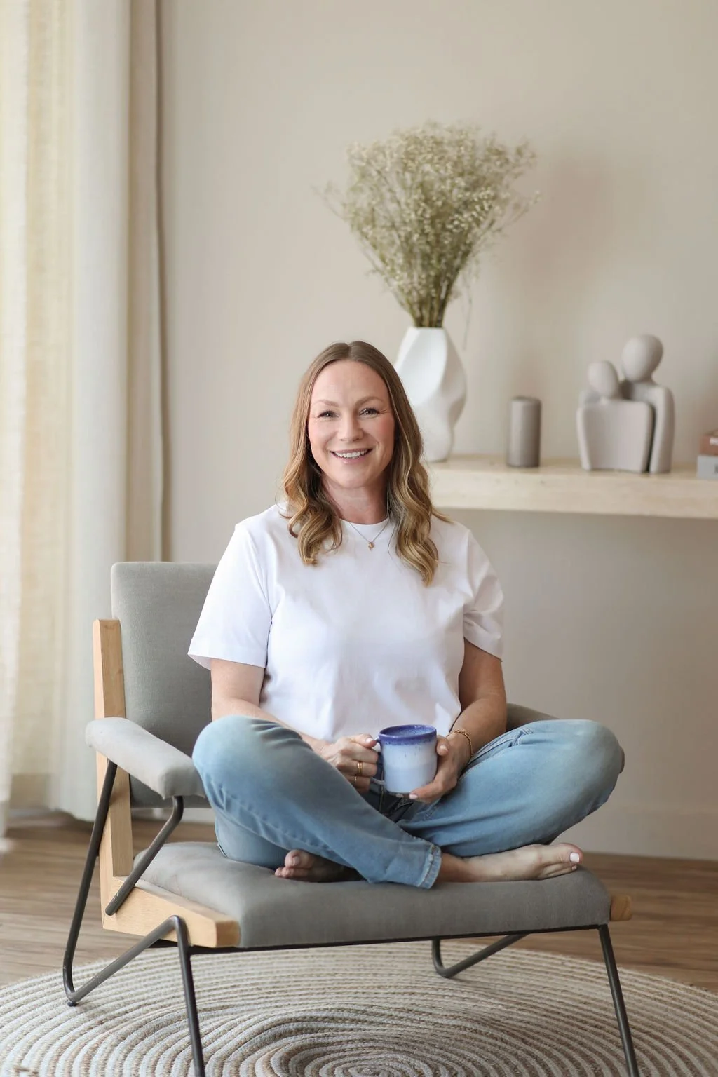 A woman sitting cross-legged on a modern chair, smiling and holding a mug, in a cozy, well-lit room with minimalistic decor and neutral tones.