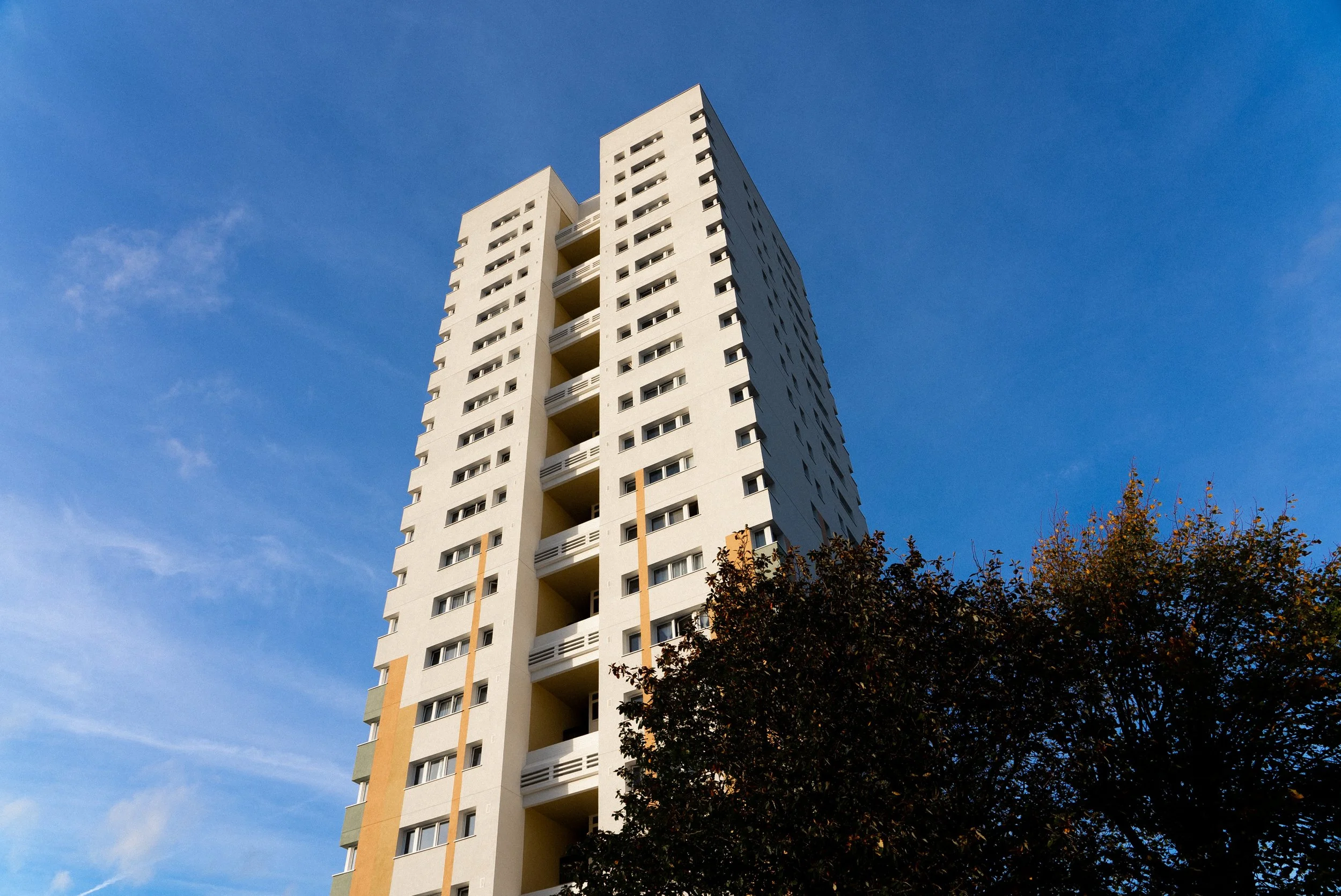 A tall white residential building with multiple balconies and windows under a clear blue sky, with a tree at the bottom.