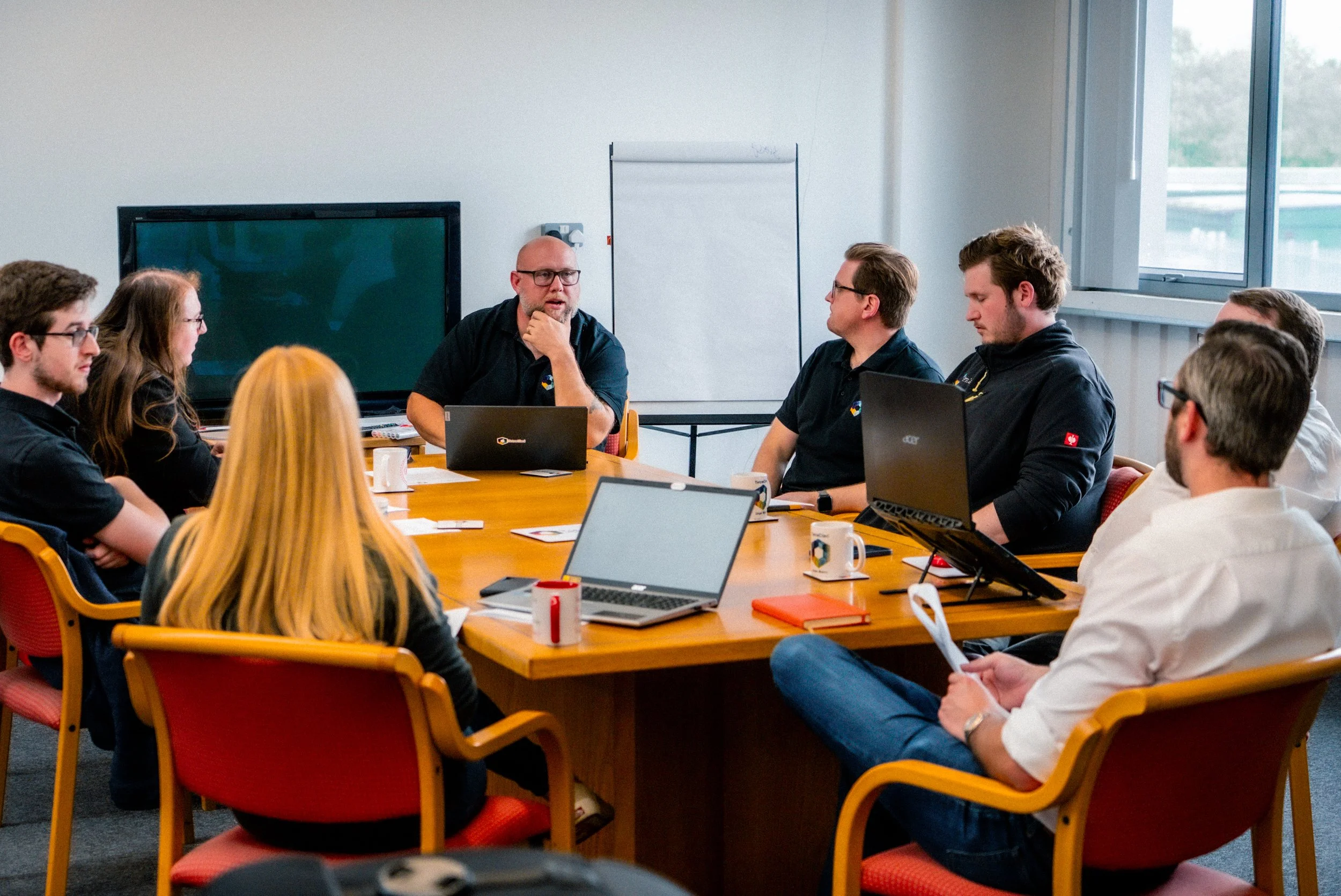 A group of nine people sitting around a conference table in a business meeting, with laptops, coffee mugs, and documents, while the main speaker talks in front of a whiteboard and screen.