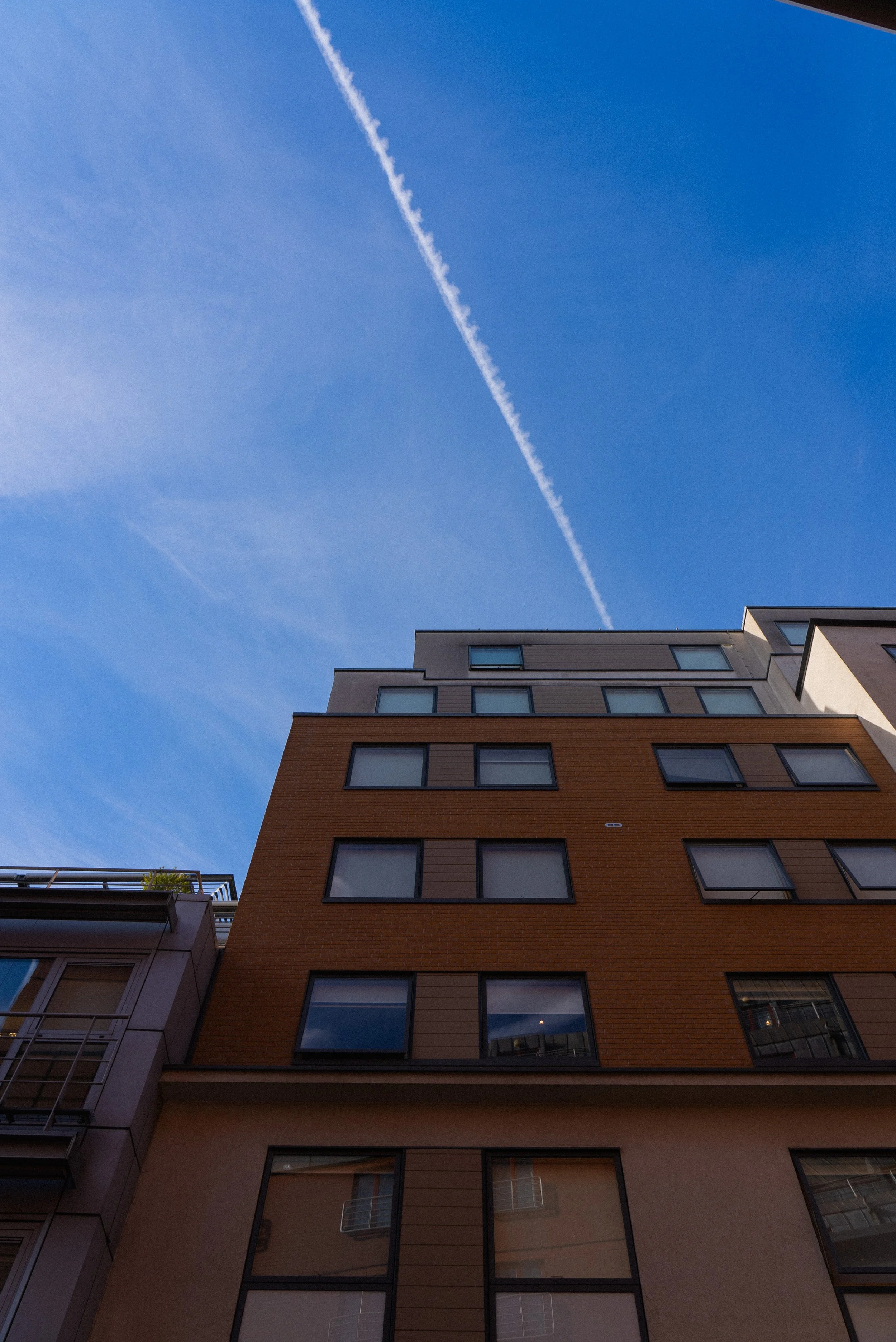 Low angle view of a multi-story residential building with large windows against a blue sky with a white contrail.