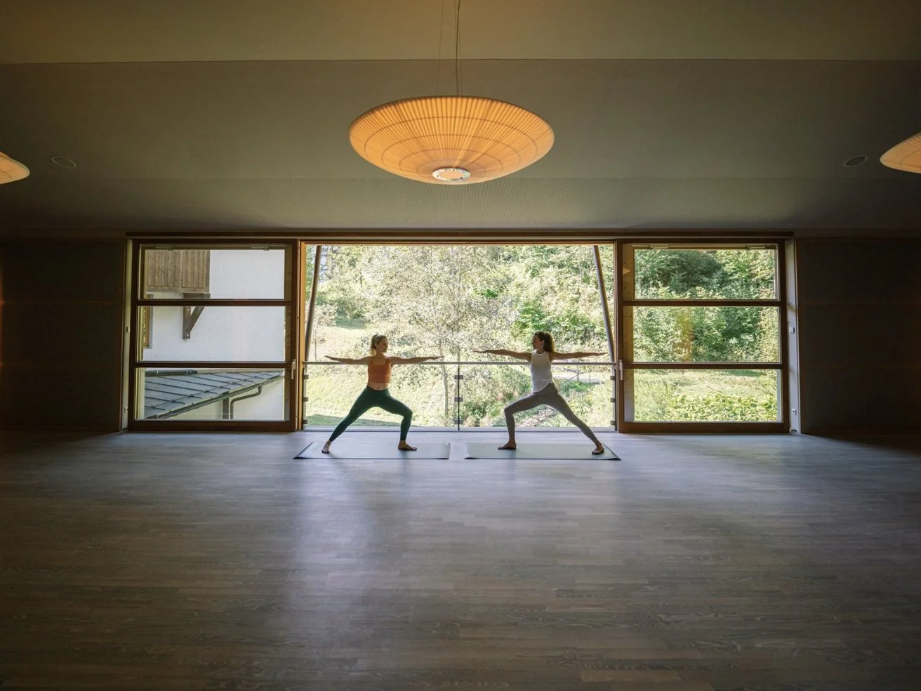Zwei Frauen machen Yoga in einem Raum mit großen Fenstern, die eine grüne Landschaft zeigen. Sie stehen sich gegenüber in einer Yoga-Pose auf Yogamatten.