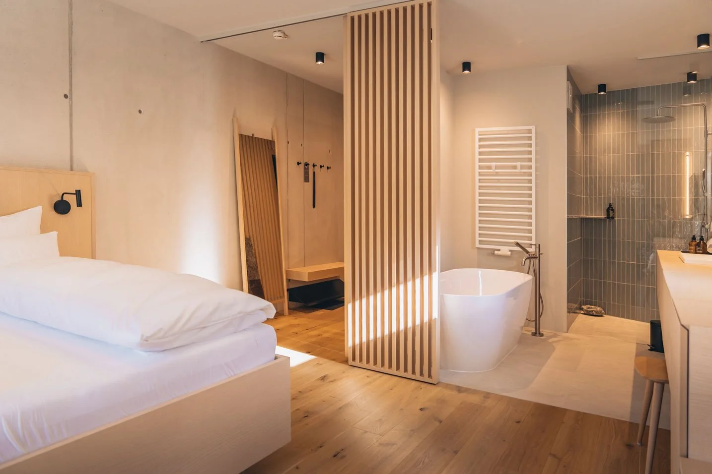 Minimalistisches Hotelzimmer mit Holzboden, einem Bett, freistehender Badewanne, Duschbereich und großen Spiegel. Moderne, schlichte Dekoration mit hellen Tönen.