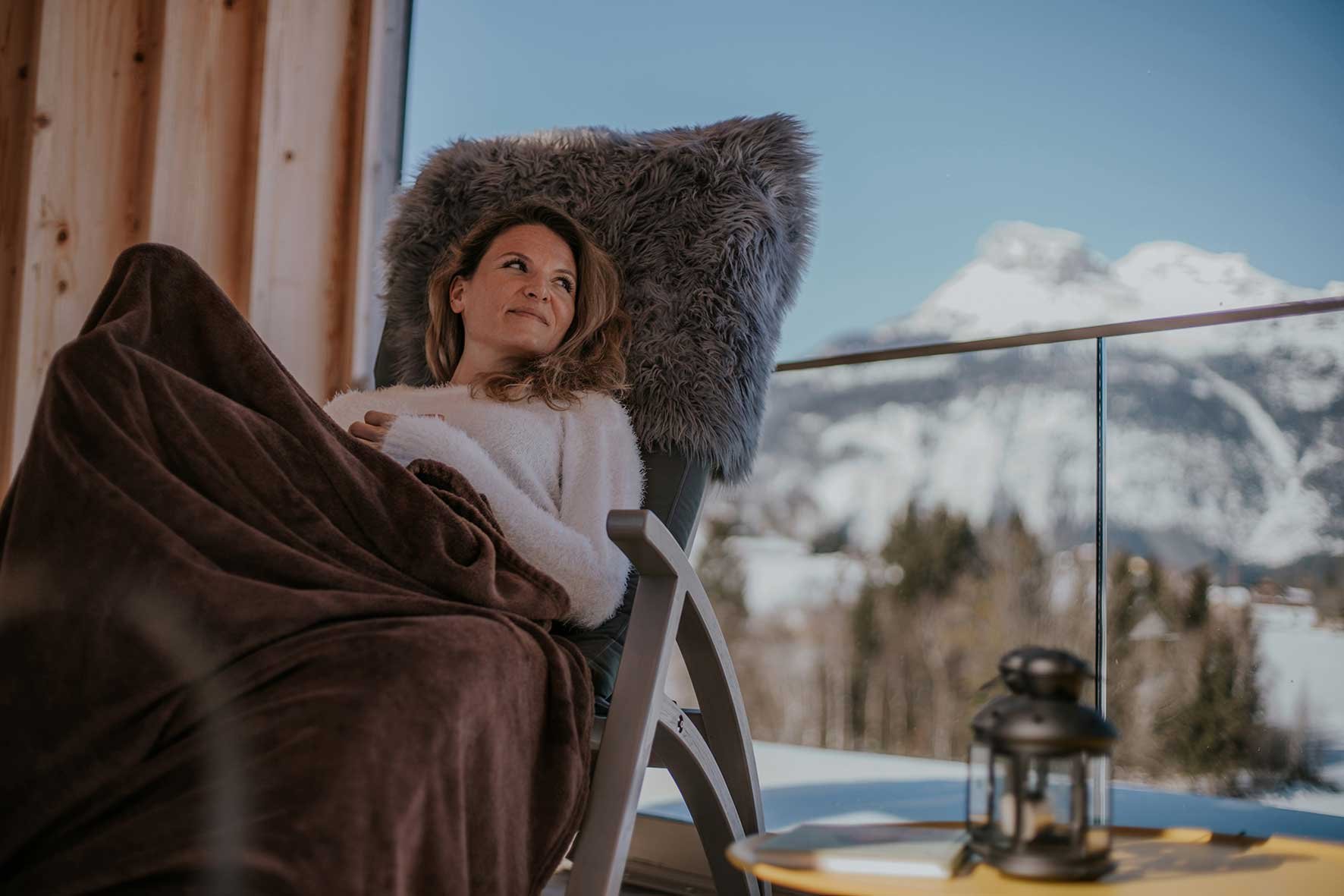 Frau liegt auf einer Couch im Chalet, schaut nach draußen auf schneebedeckte Berge und genießt die winterliche Aussicht.