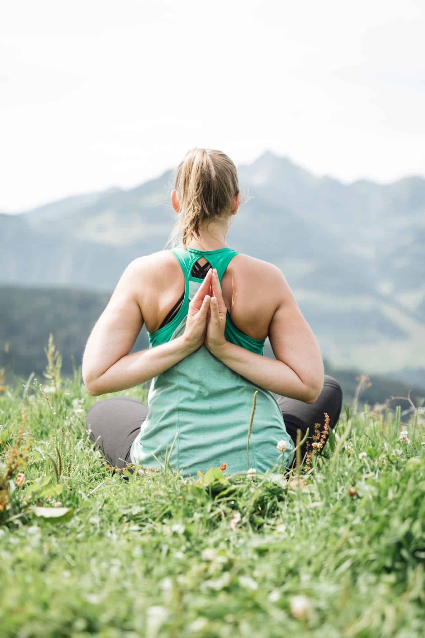 Frau macht Yoga in der Natur mit Blick auf Berge und Wiese, sitzt im Lotussitz mit wiederholender Handhaltung hinter dem Rücken.