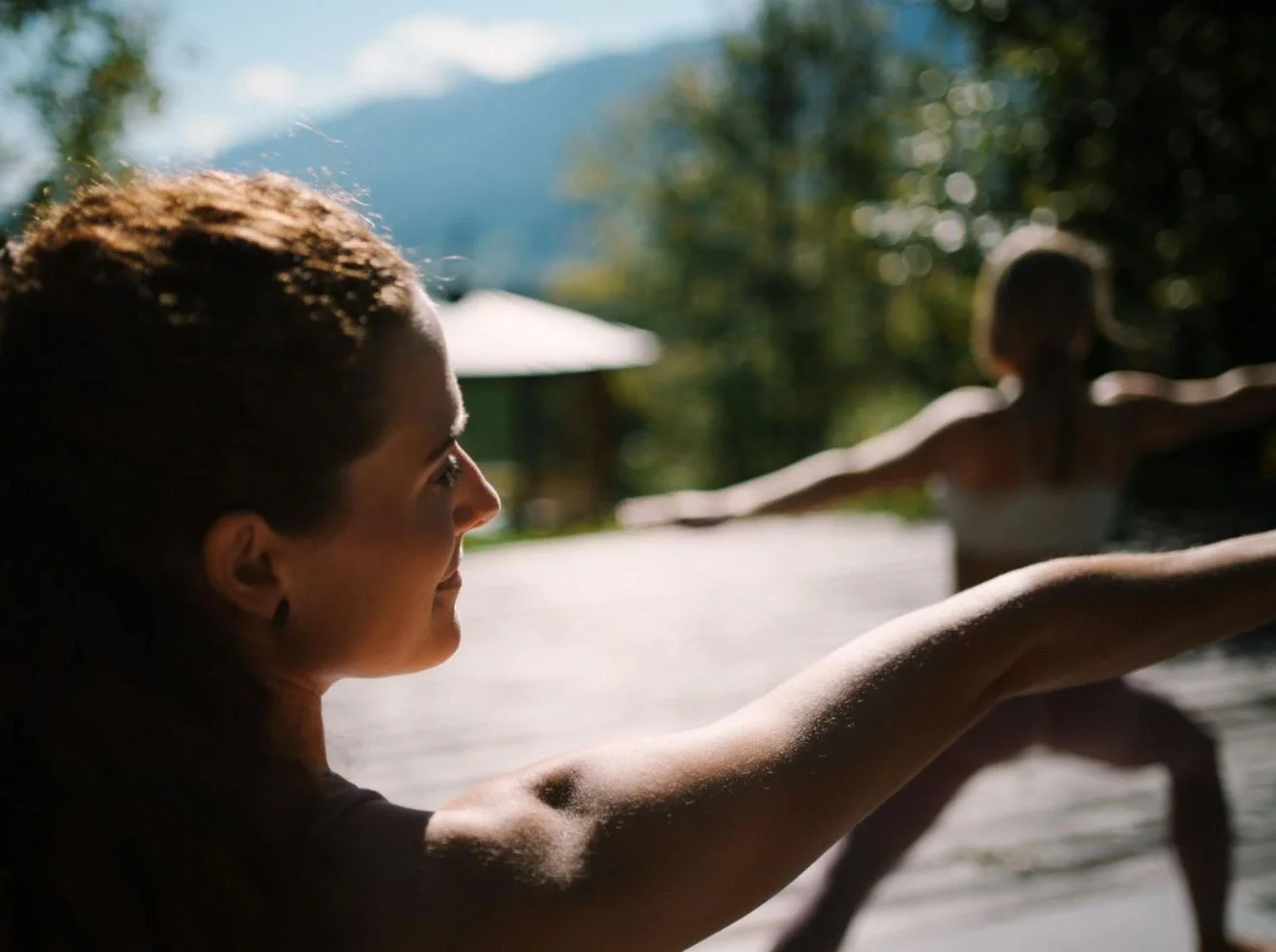 Zwei Frauen beim Yoga im Freien, eine im Vordergrund mit nach vorne ausgestreckten Armen, die andere im Hintergrund bei Yoga-Übung, umgeben von grüner Natur und Bäumen.