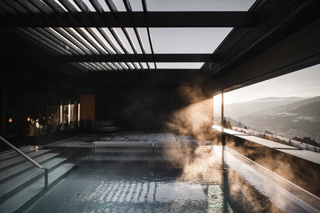 Ein modernes Spa mit einem überdachten Pool, aus dem Dampf steigt. Die offene Seite zeigt einen Blick auf eine Berglandschaft bei Sonnenuntergang.