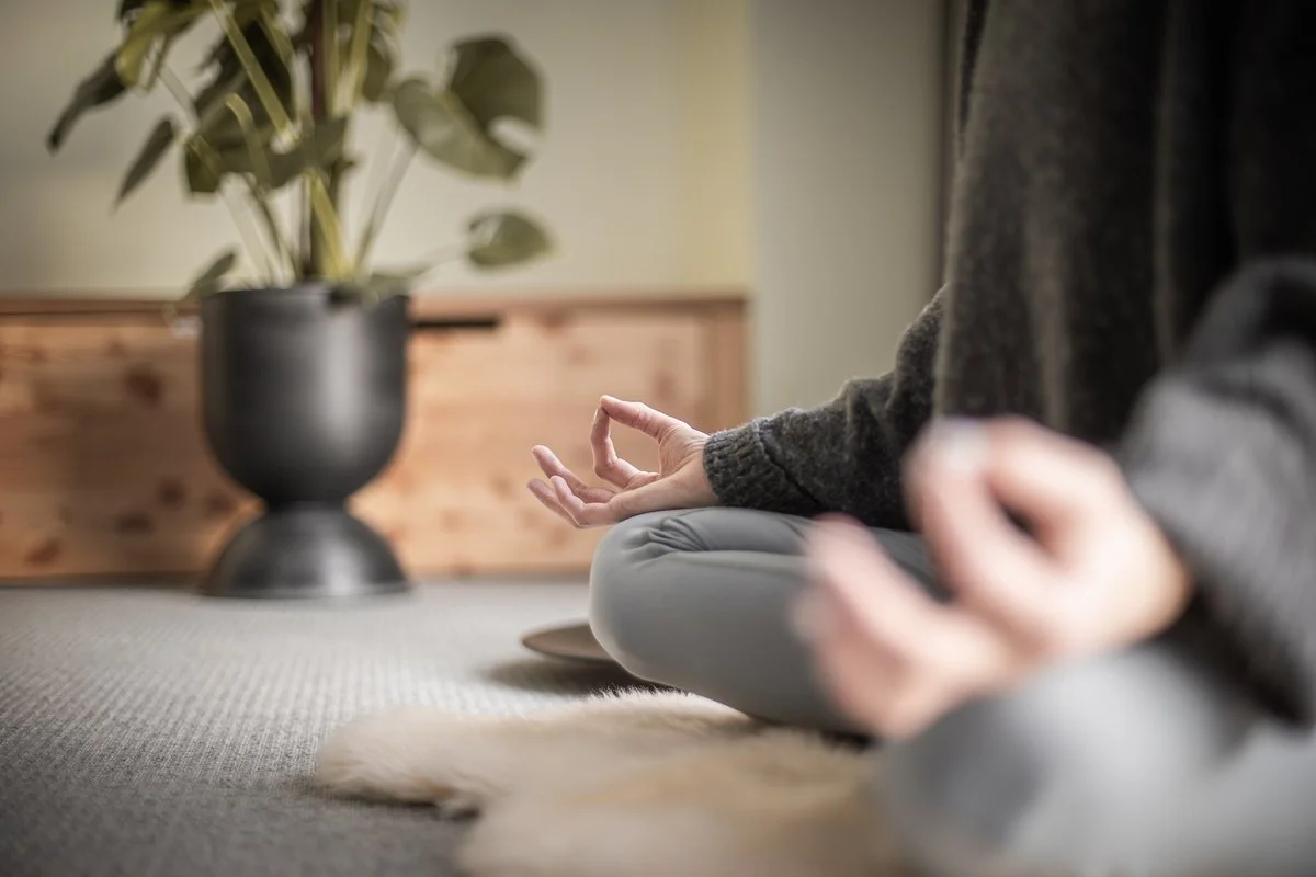 Person in sitzender Meditationspose auf einem Teppich, in einem Raum mit großem schwarzen Blumentopf und Pflanzen