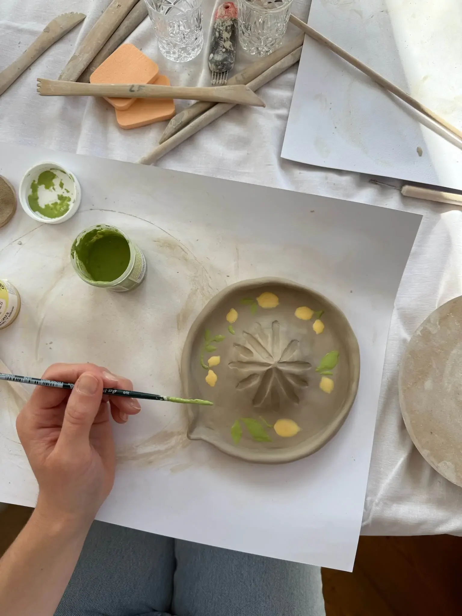 Eine Person bemalt eine Keramikschale mit grüner und gelber Farbe, während der Tisch mit Malutensilien, Gläsern und anderen Gegenständen gedeckt ist.