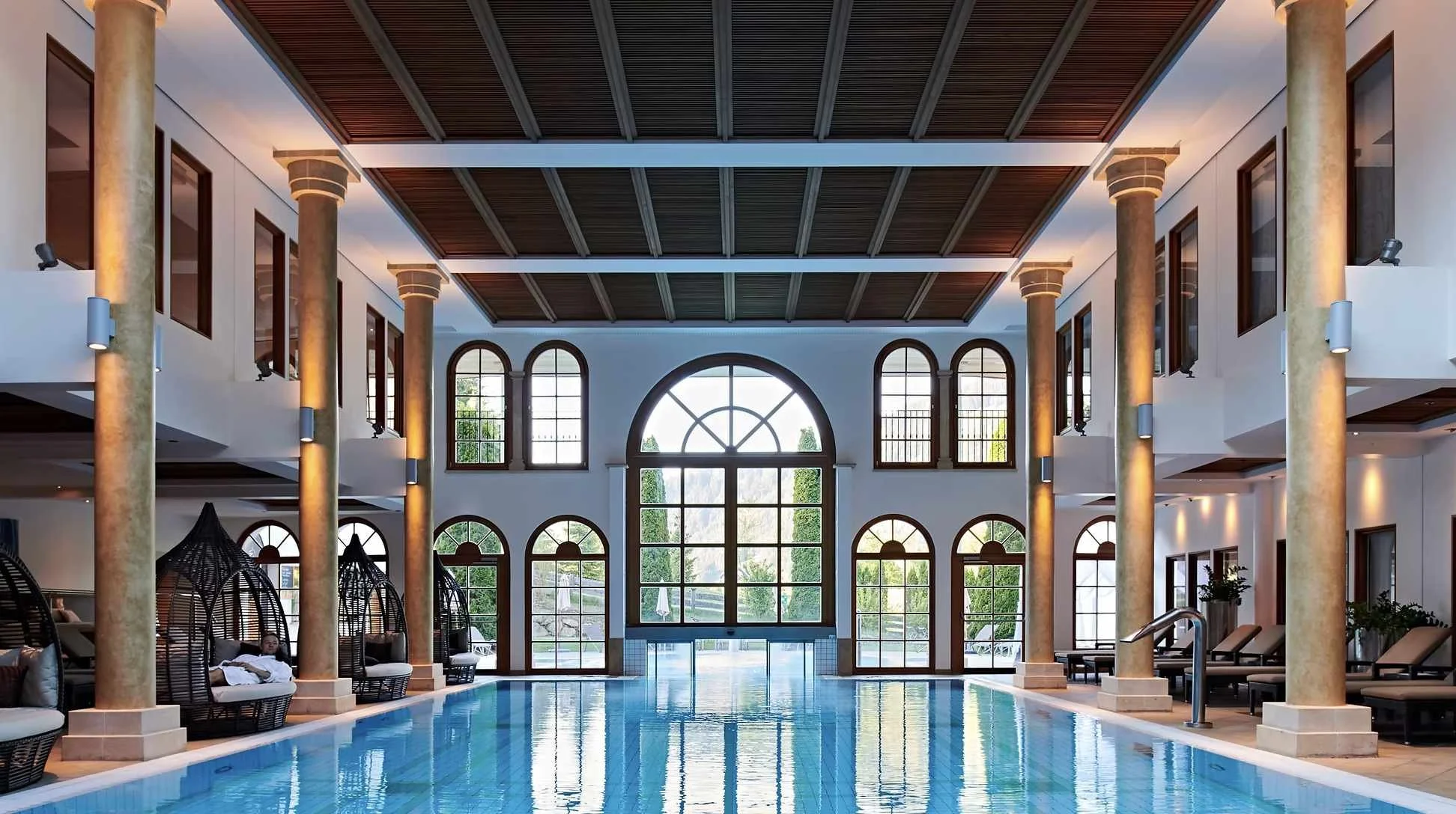 Innenraum eines luxuriösen Schwimmbads mit hohen Fenstern, goldenen Säulen und warmem Licht.