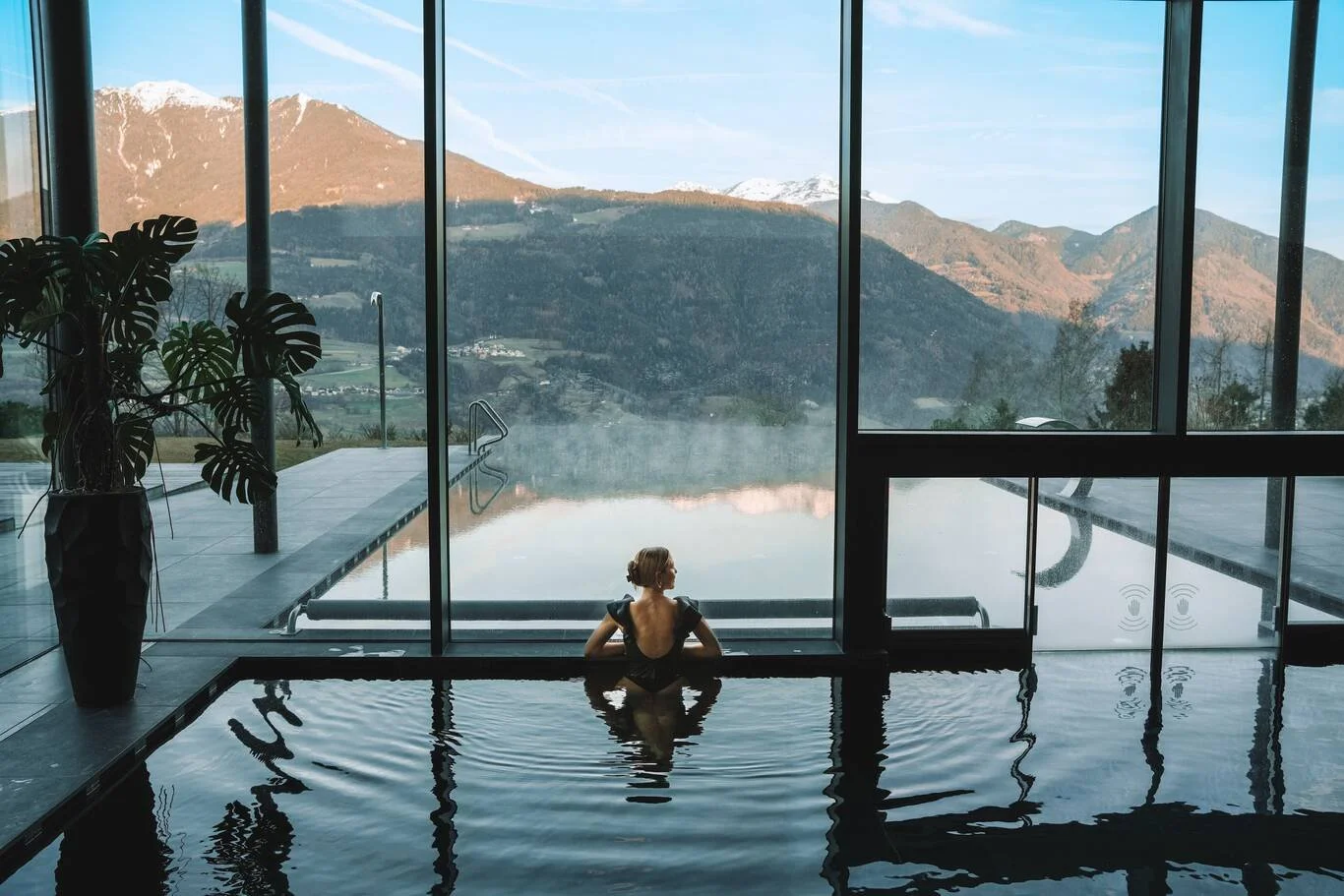 Frau entspannt im Innenpool mit Blick auf Berge und Himmel durch große Fenster